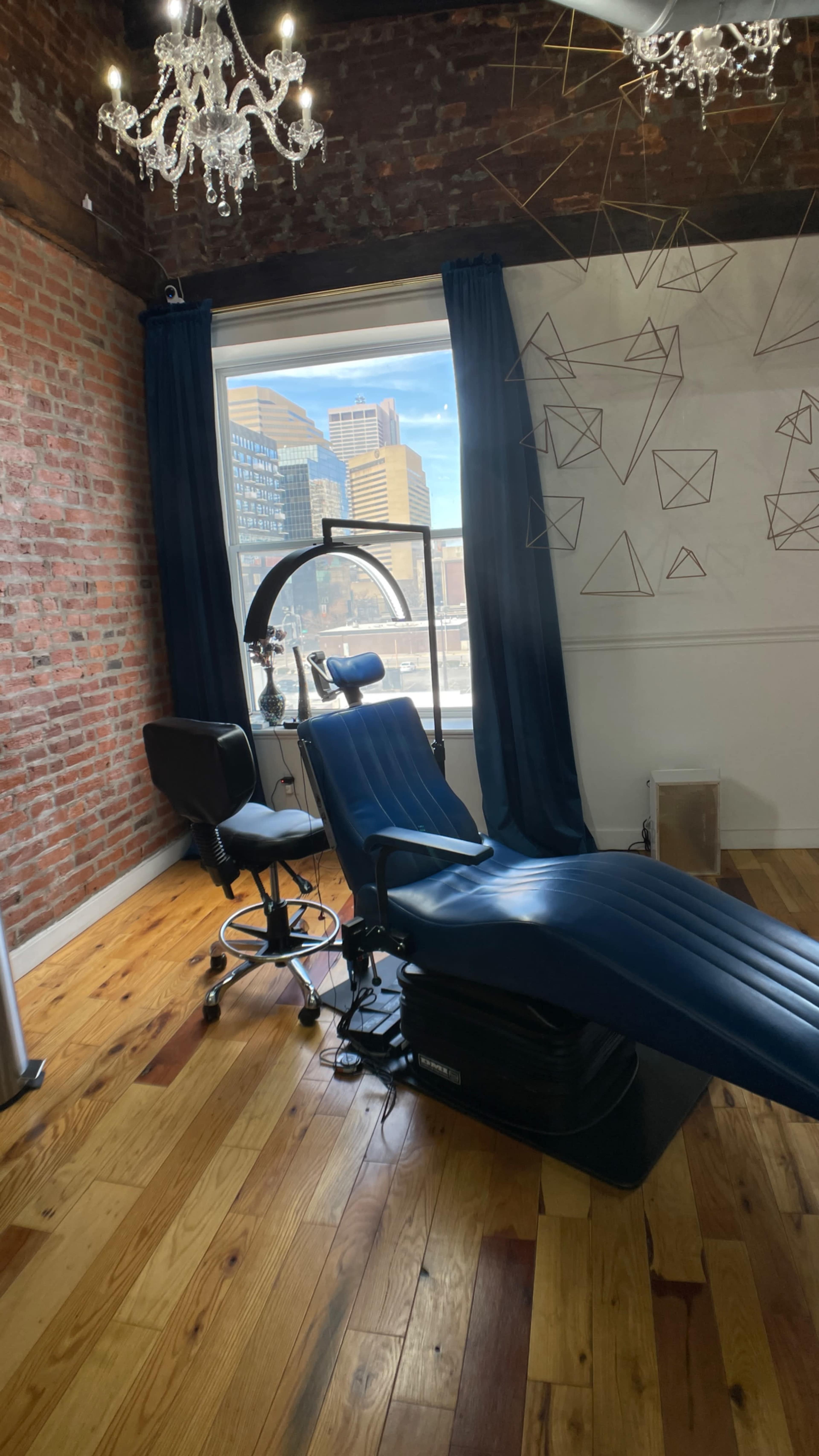 A modern room with exposed brick walls, a large window providing city views, a blue treatment chair, and a styling chair nearby.