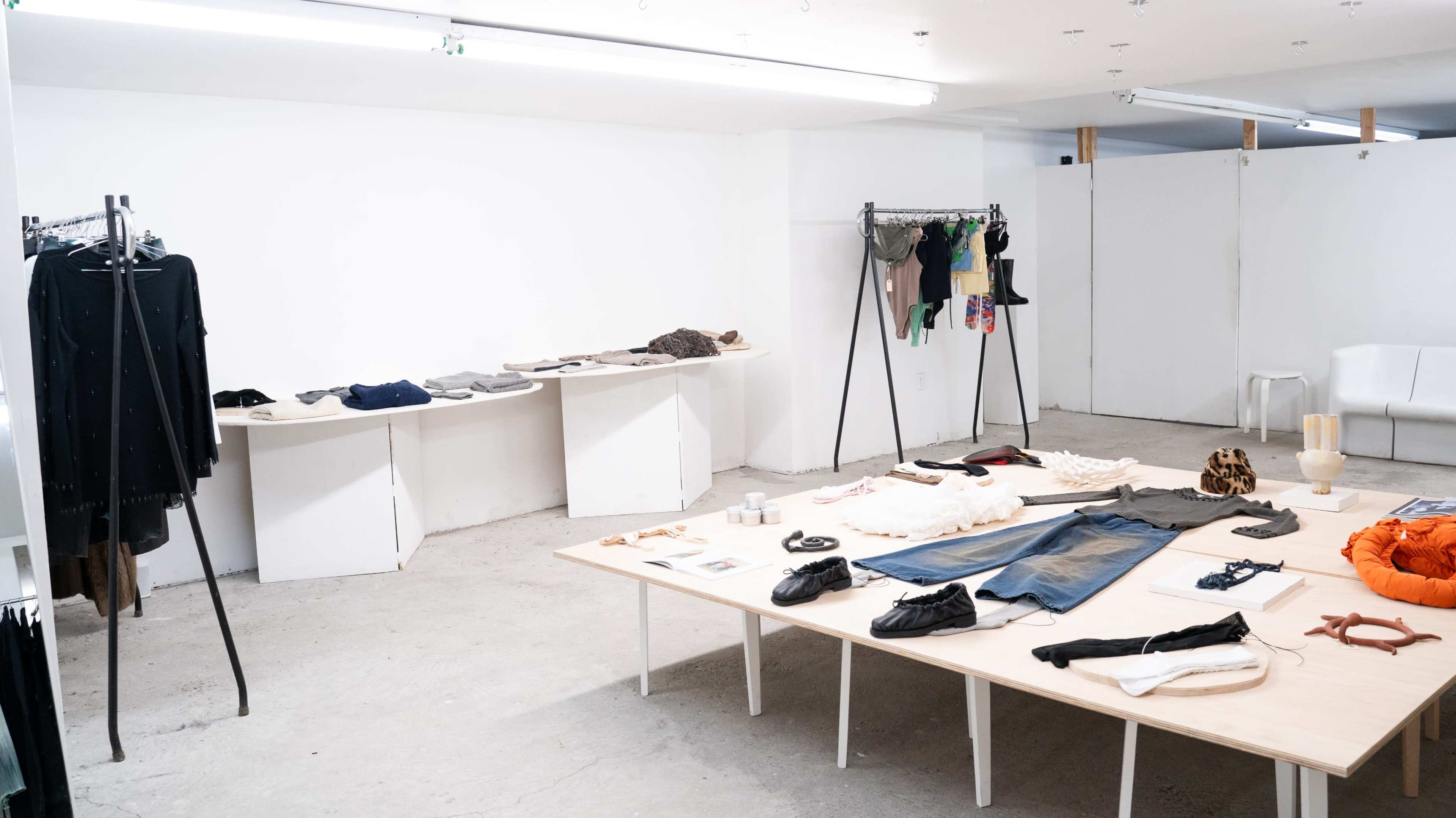 A minimalist clothing store interior with garments displayed on tables and racks against a plain white wall.