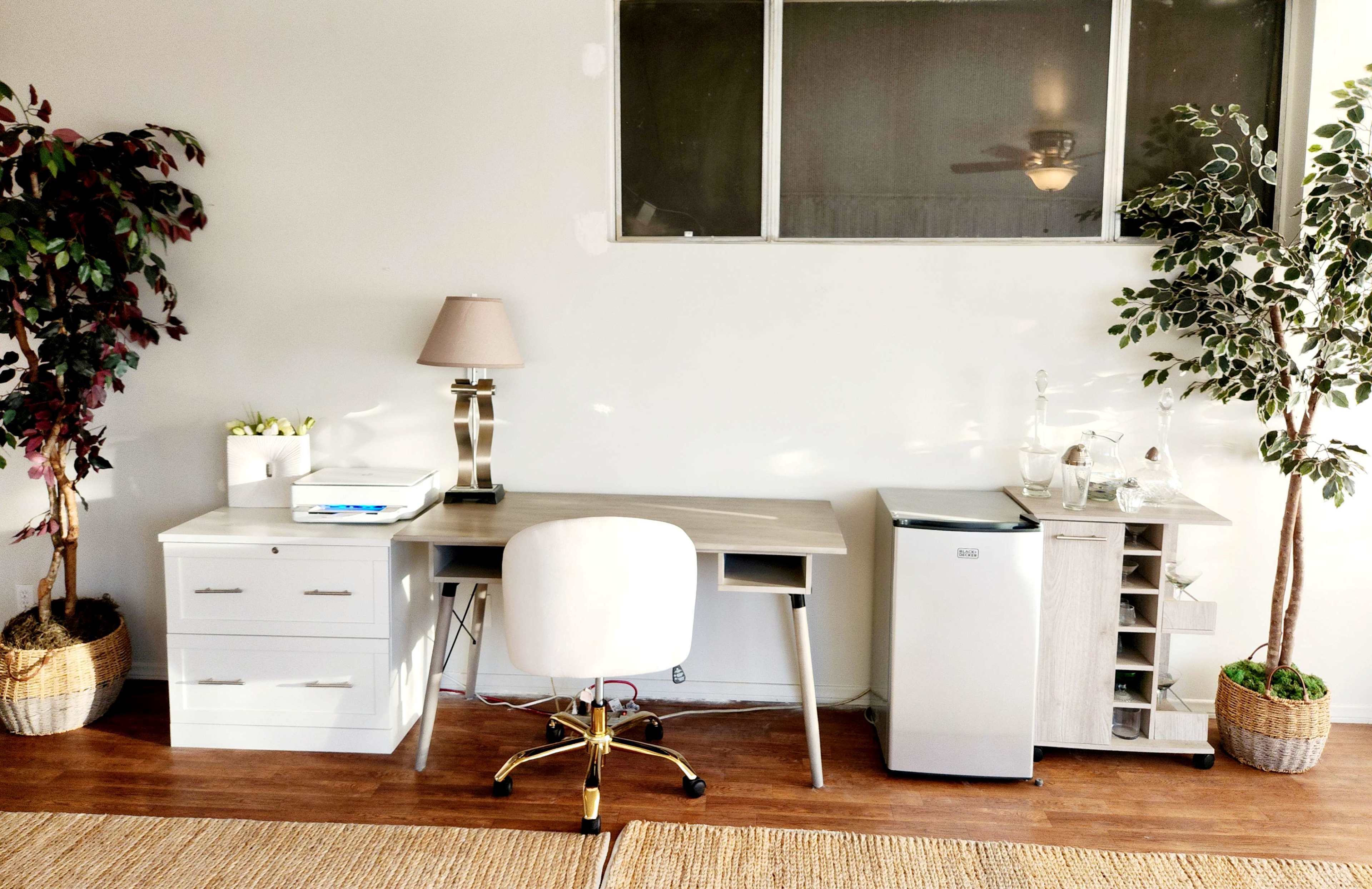 A minimalist home office setup features a wooden desk with a chair, a white filing cabinet, a small refrigerator, and decorative plants in the corners.