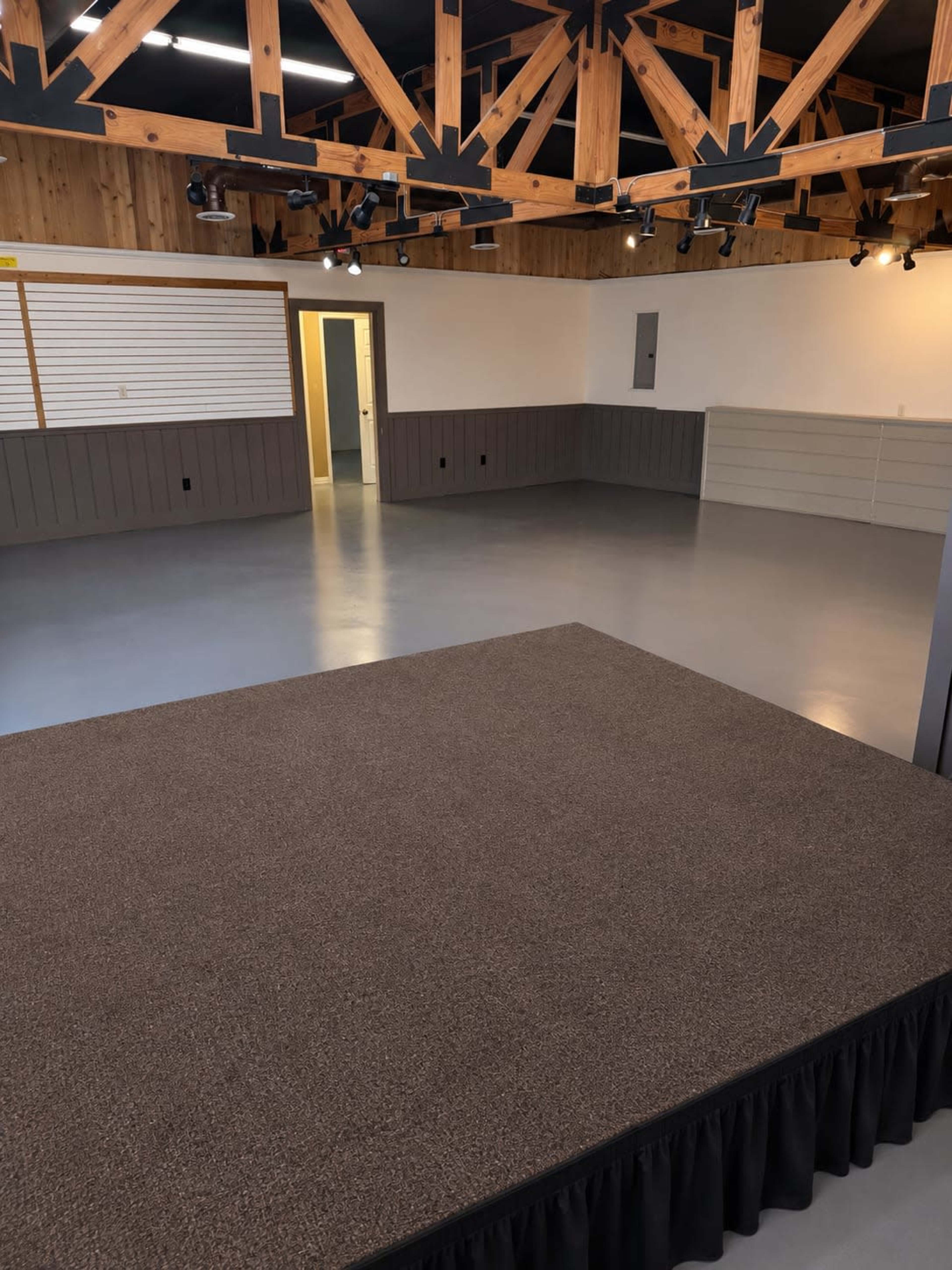 The image shows an empty room with wooden beams on the ceiling, a raised carpeted stage, and a simple wall design.