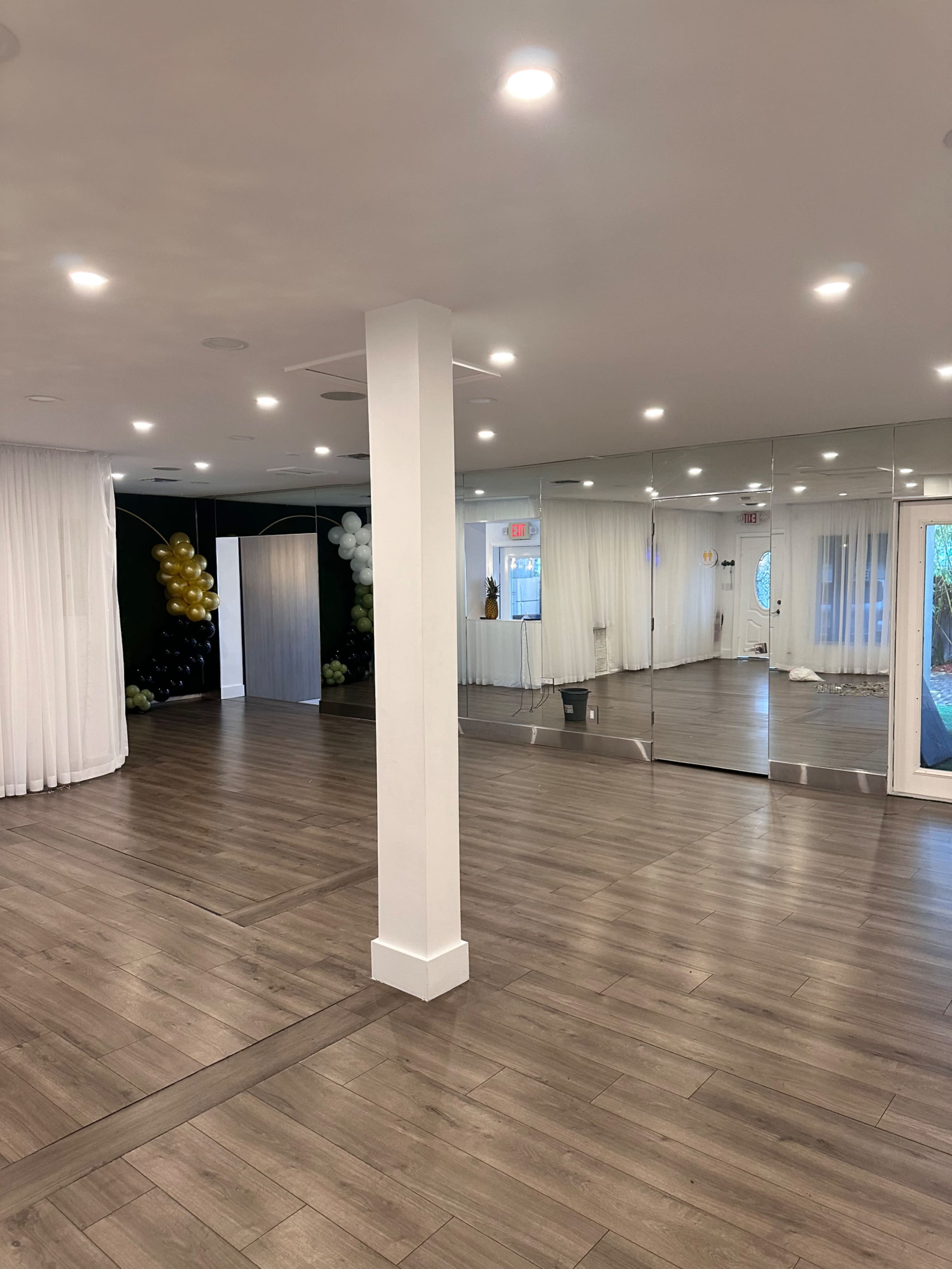 The image shows a spacious indoor area with wooden flooring and mirrored walls, featuring white curtains and decorative balloons in one corner.