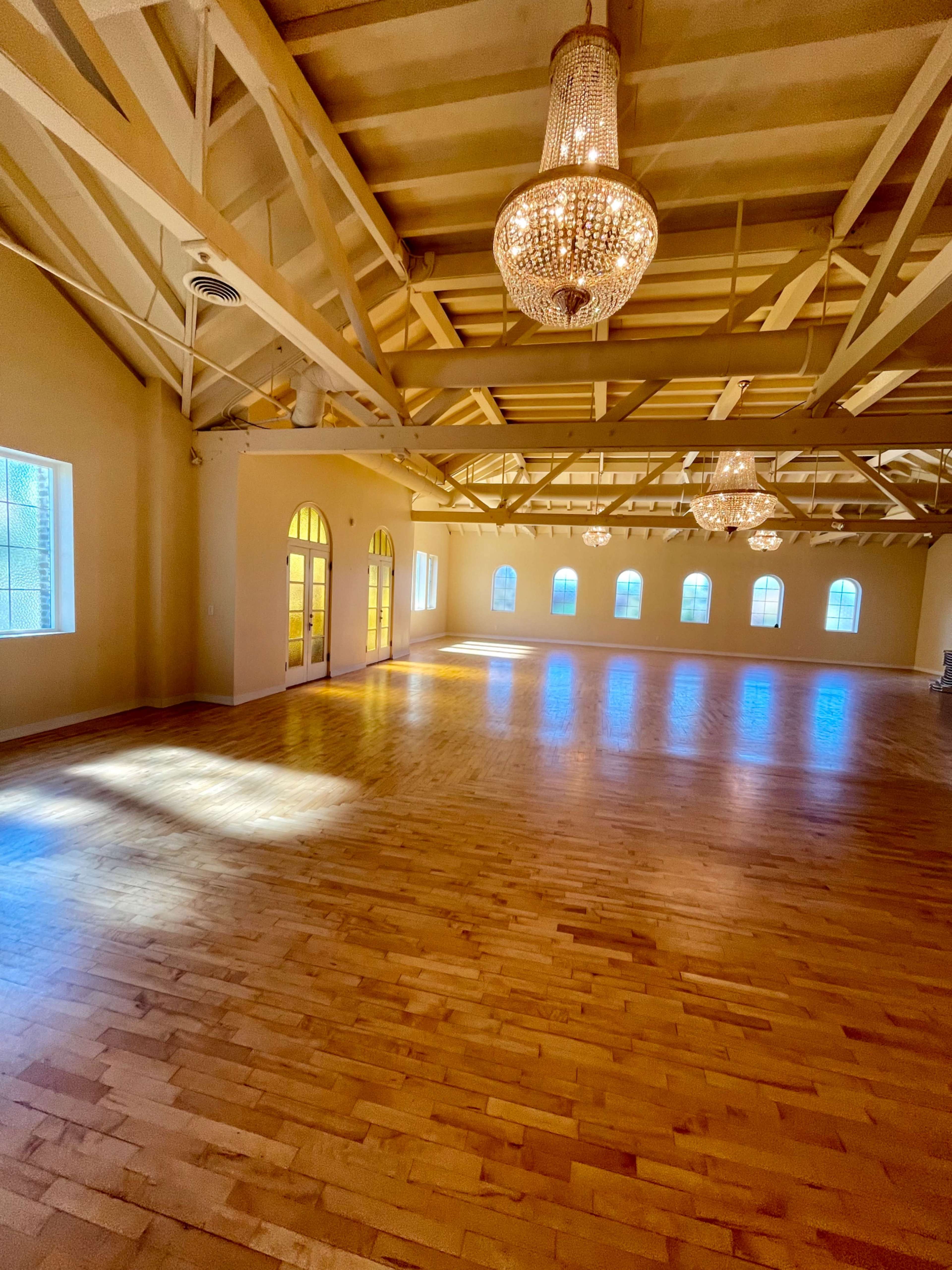 The image shows a spacious, well-lit room with wooden floors, high ceilings, and large windows, adorned with three chandeliers.