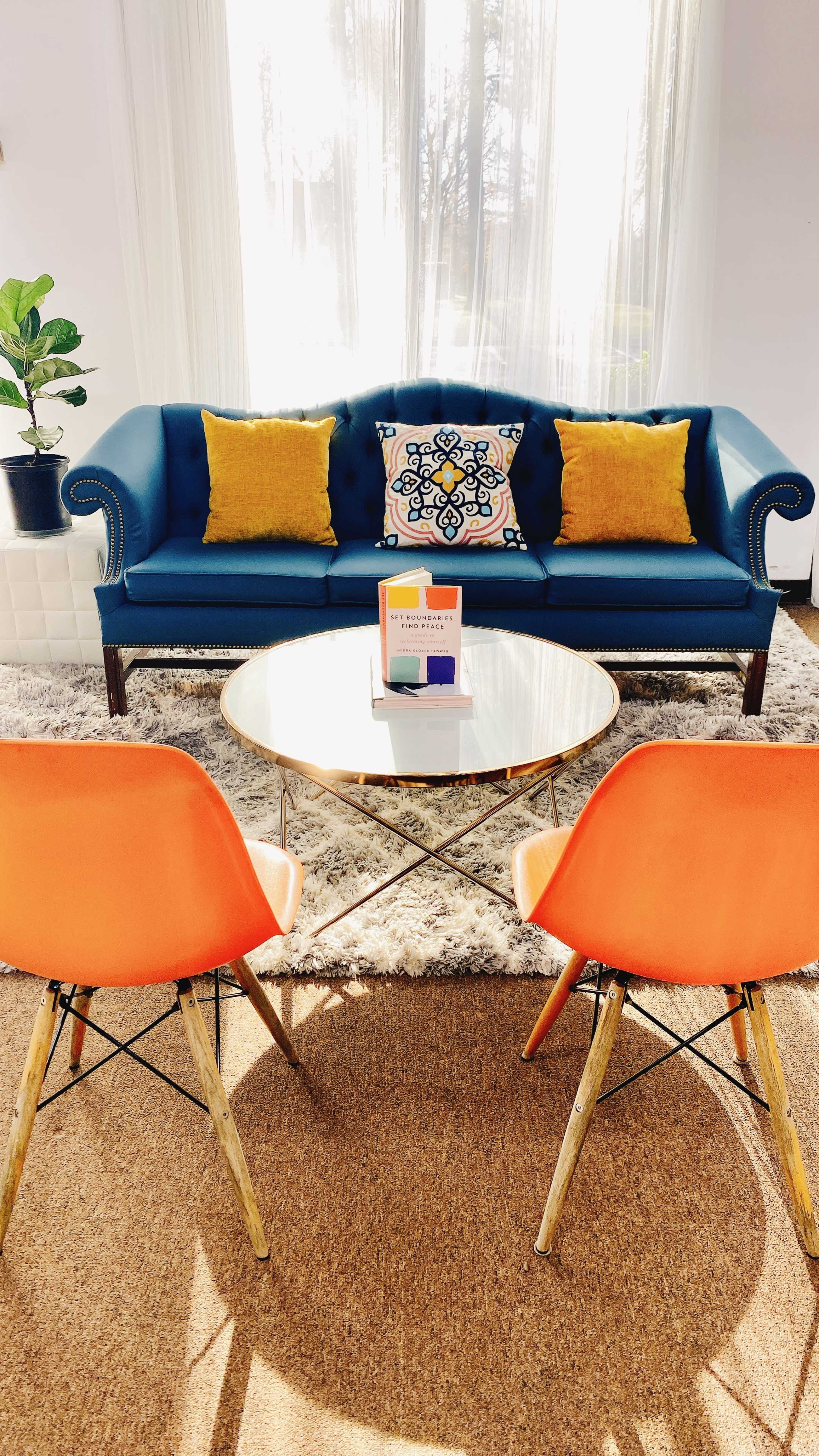 A blue couch with yellow pillows is positioned in front of a round gold coffee table, flanked by two orange chairs on a patterned rug in a brightly lit room.