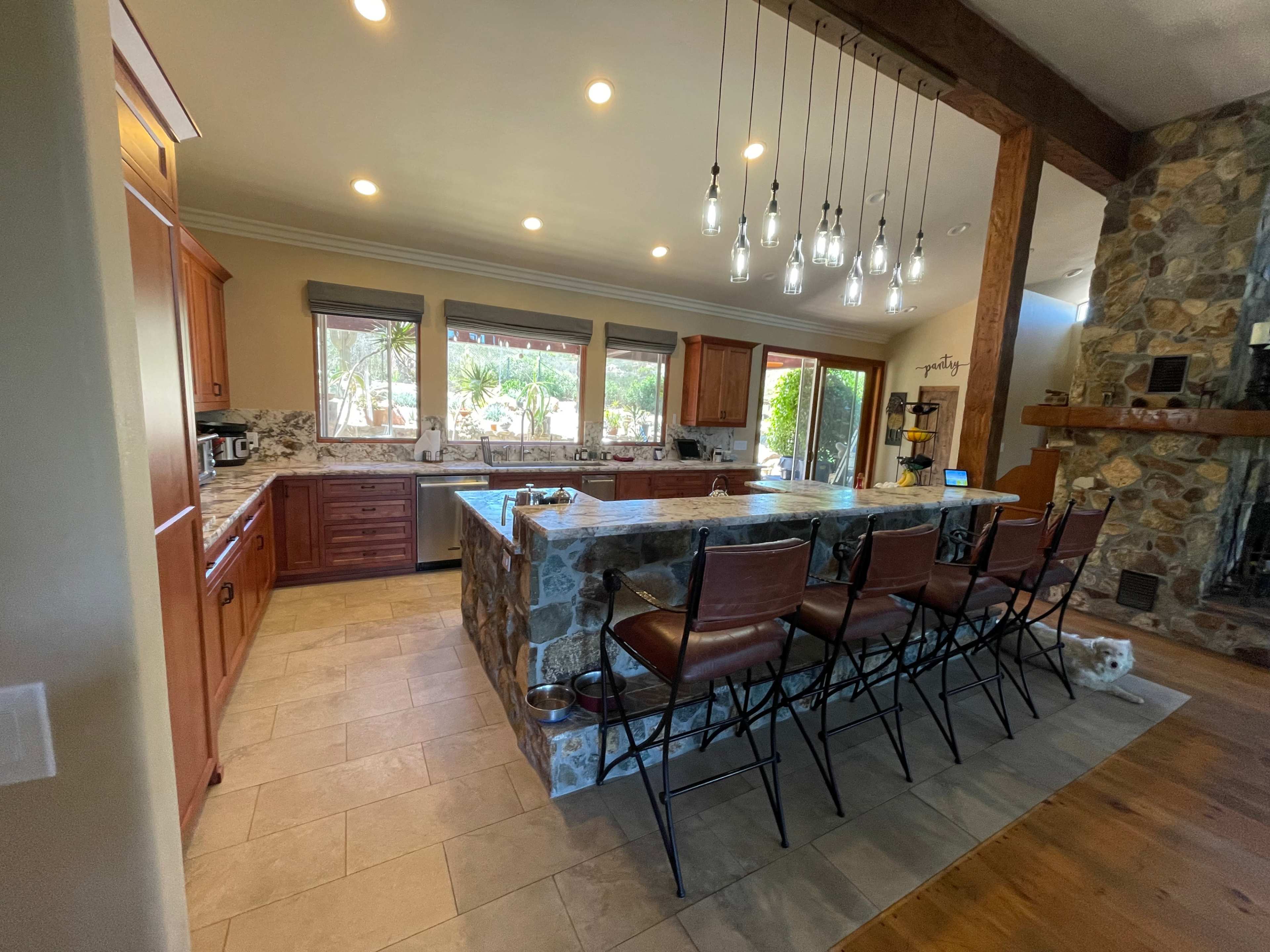 The image shows a spacious kitchen with wooden cabinets, a stone island, and a seating area featuring barstools, illuminated by pendant lights.