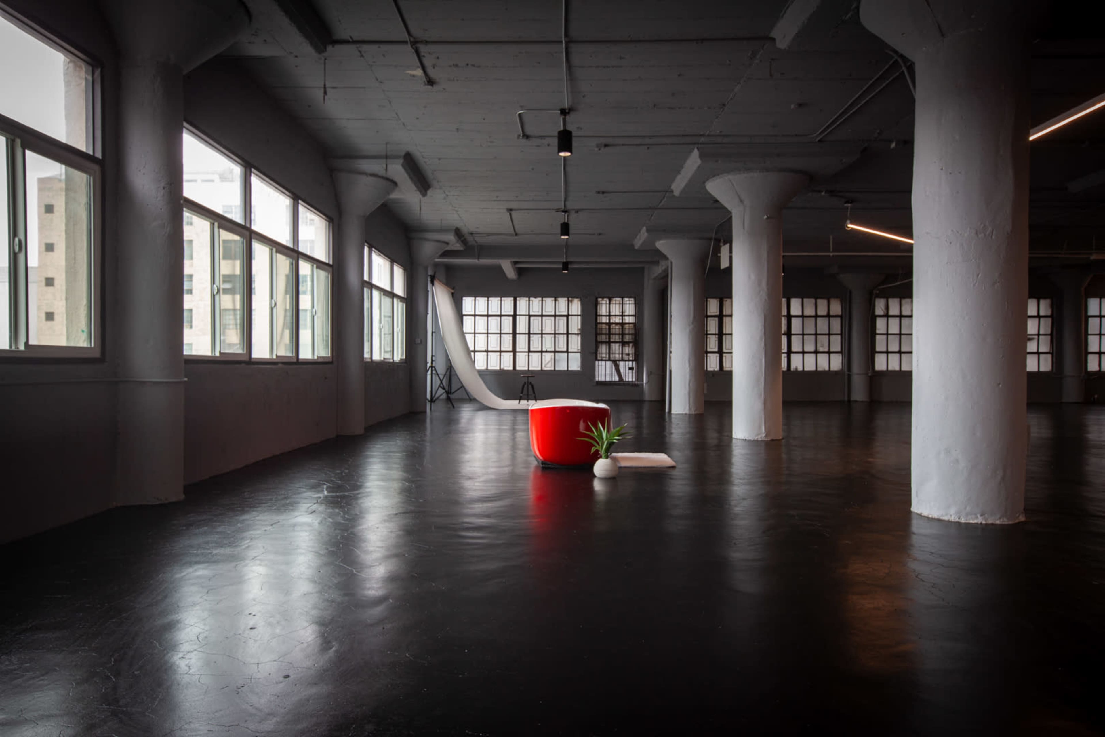 The image shows a spacious, industrial-style room with large windows, concrete pillars, and a red chair positioned on a polished black floor.