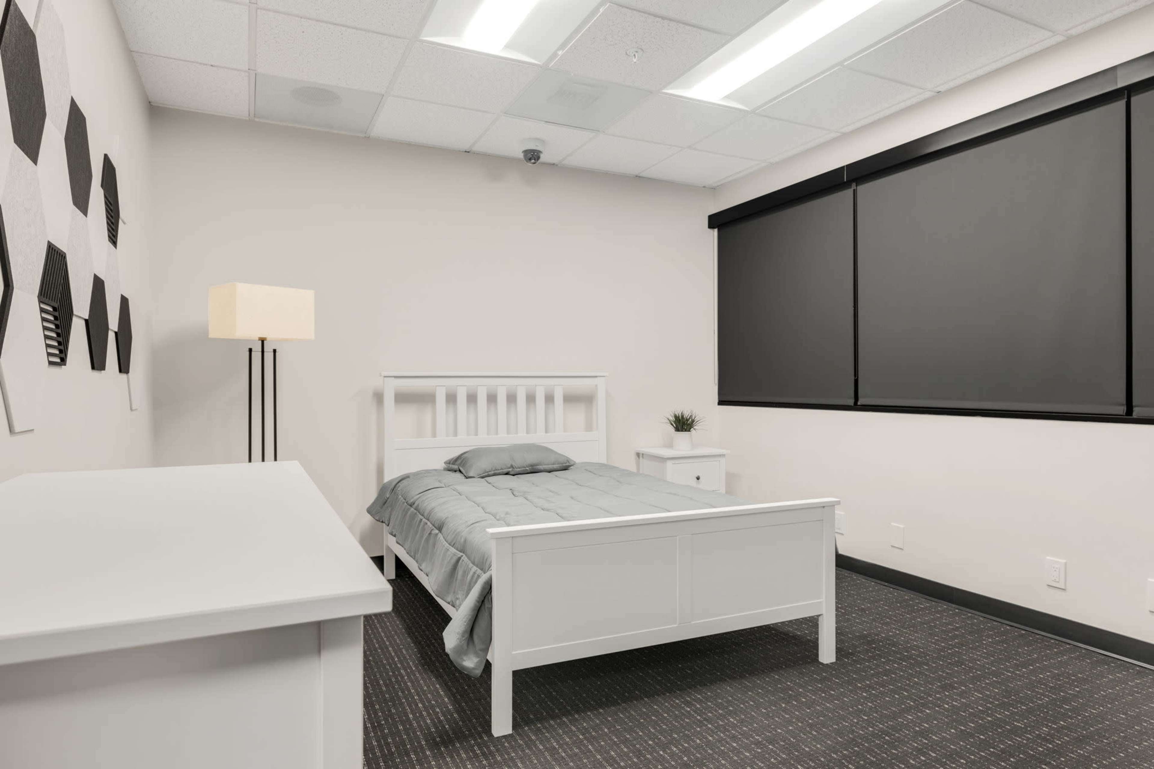 The image shows a simple bedroom with a white bed, nightstand, and a lamp, set against a light-colored wall and dark window blinds.