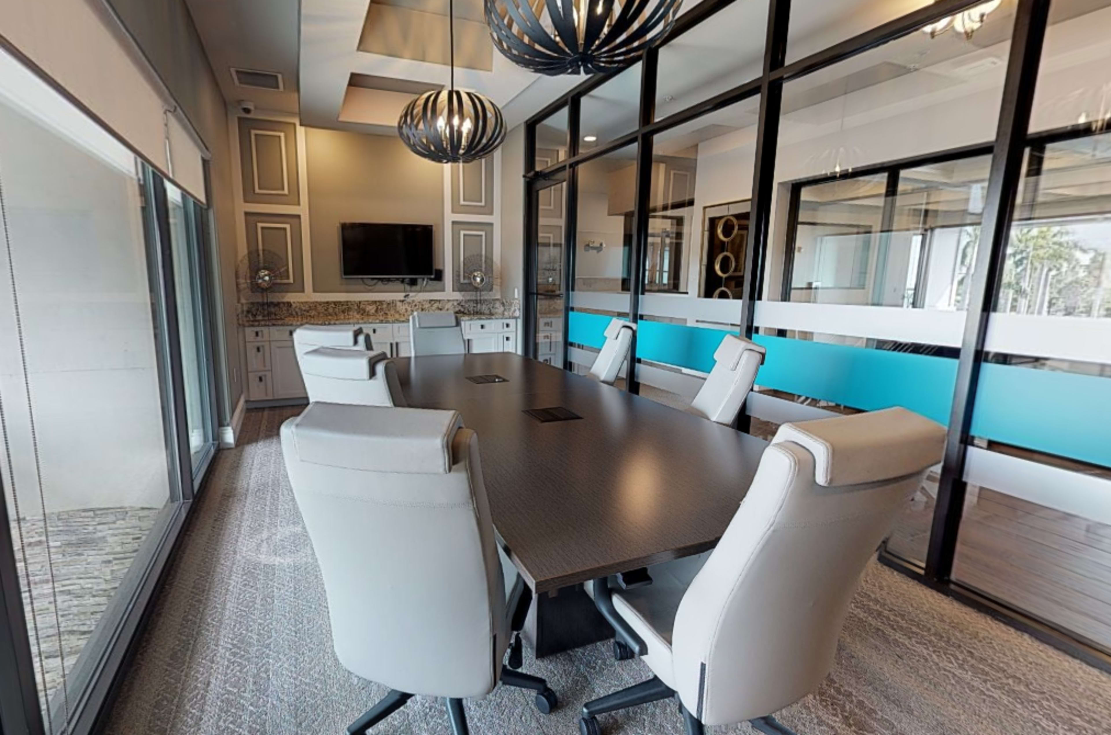 A modern conference room features a large table surrounded by white chairs, with a TV on the wall and decorative lighting fixtures above.