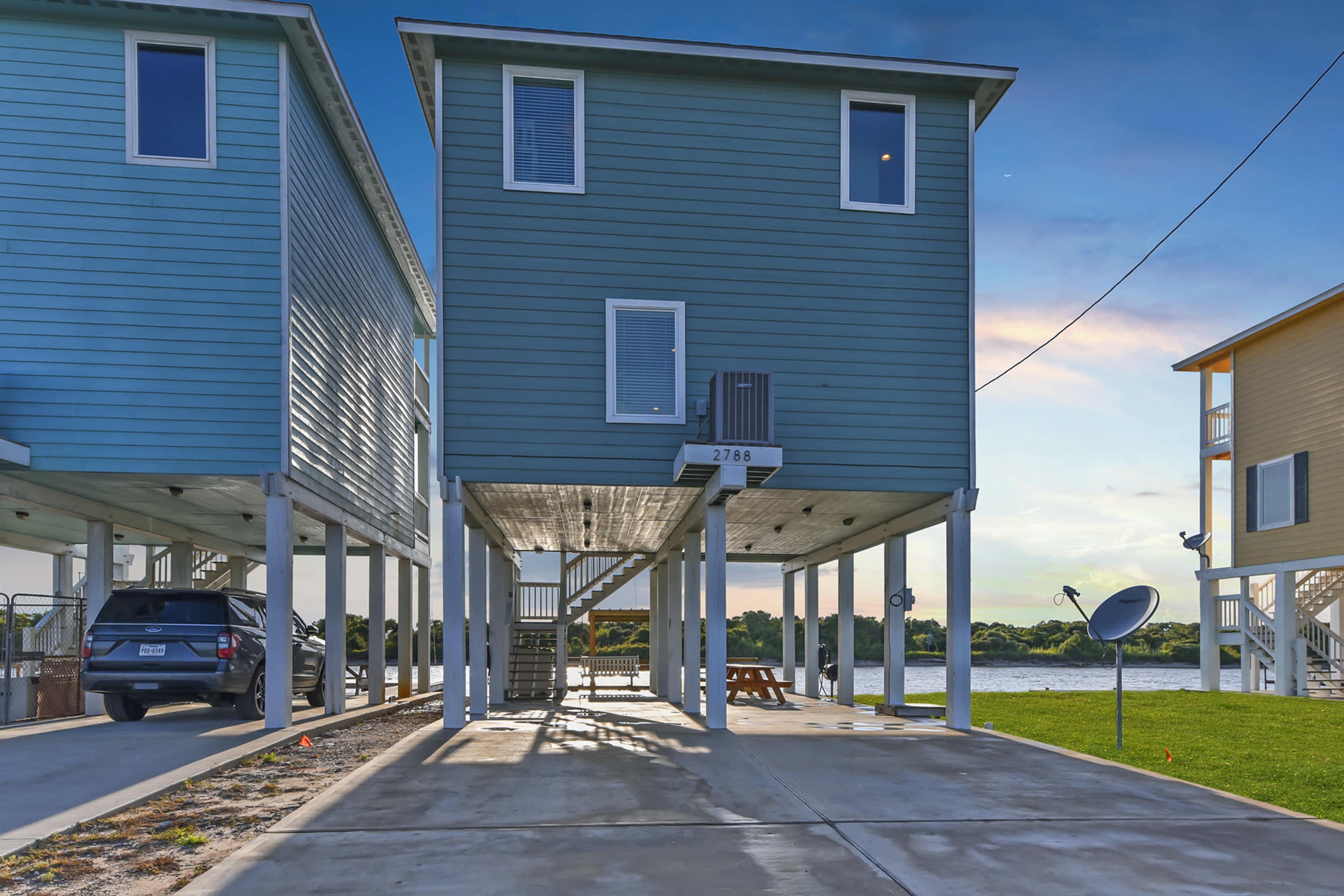 A two-story house stands on stilts near a waterfront, with an outdoor parking area underneath and satellite dishes positioned nearby.