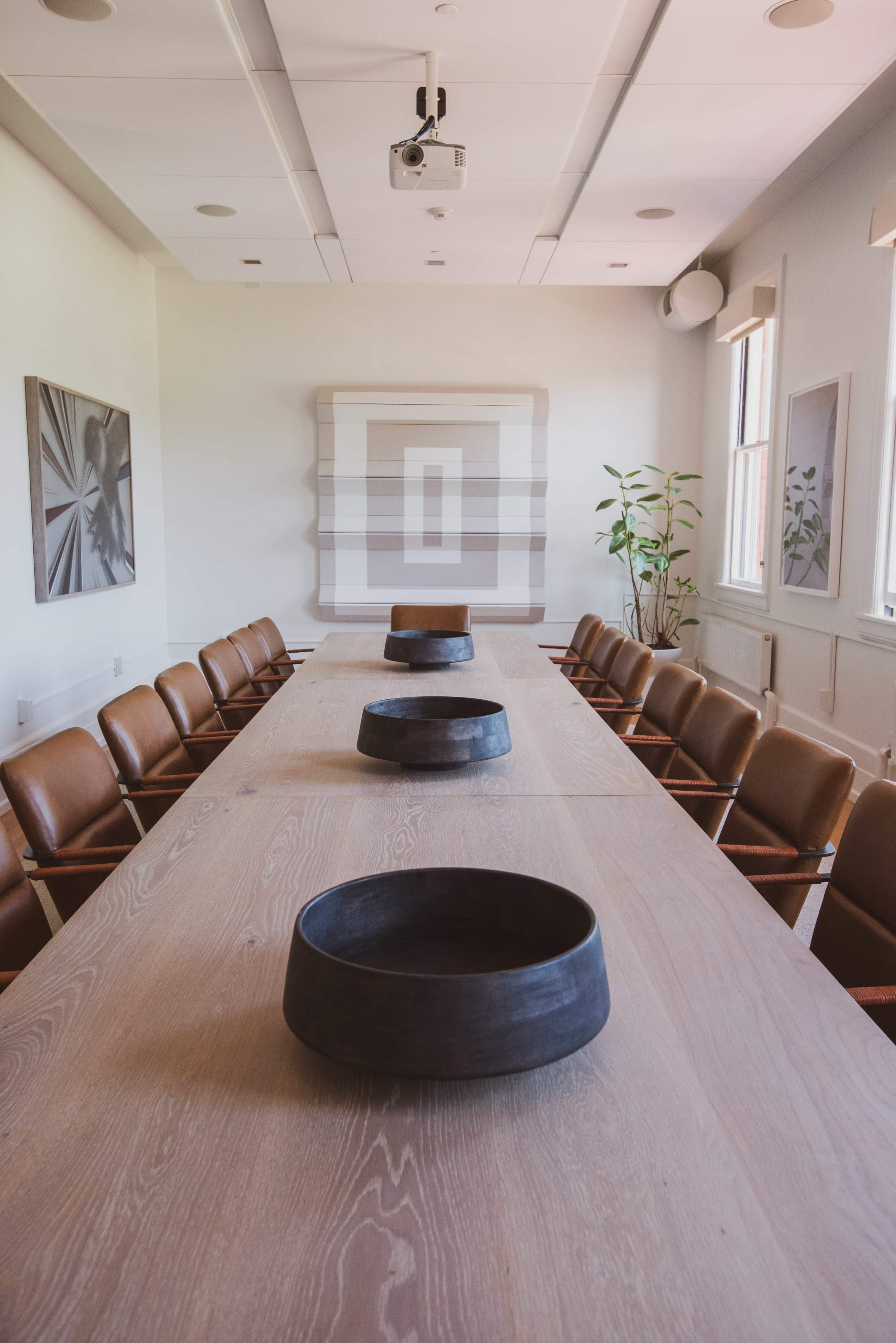 A long wooden conference table is set with dark bowls in a bright, modern meeting room featuring a projector and framed artworks on the walls.