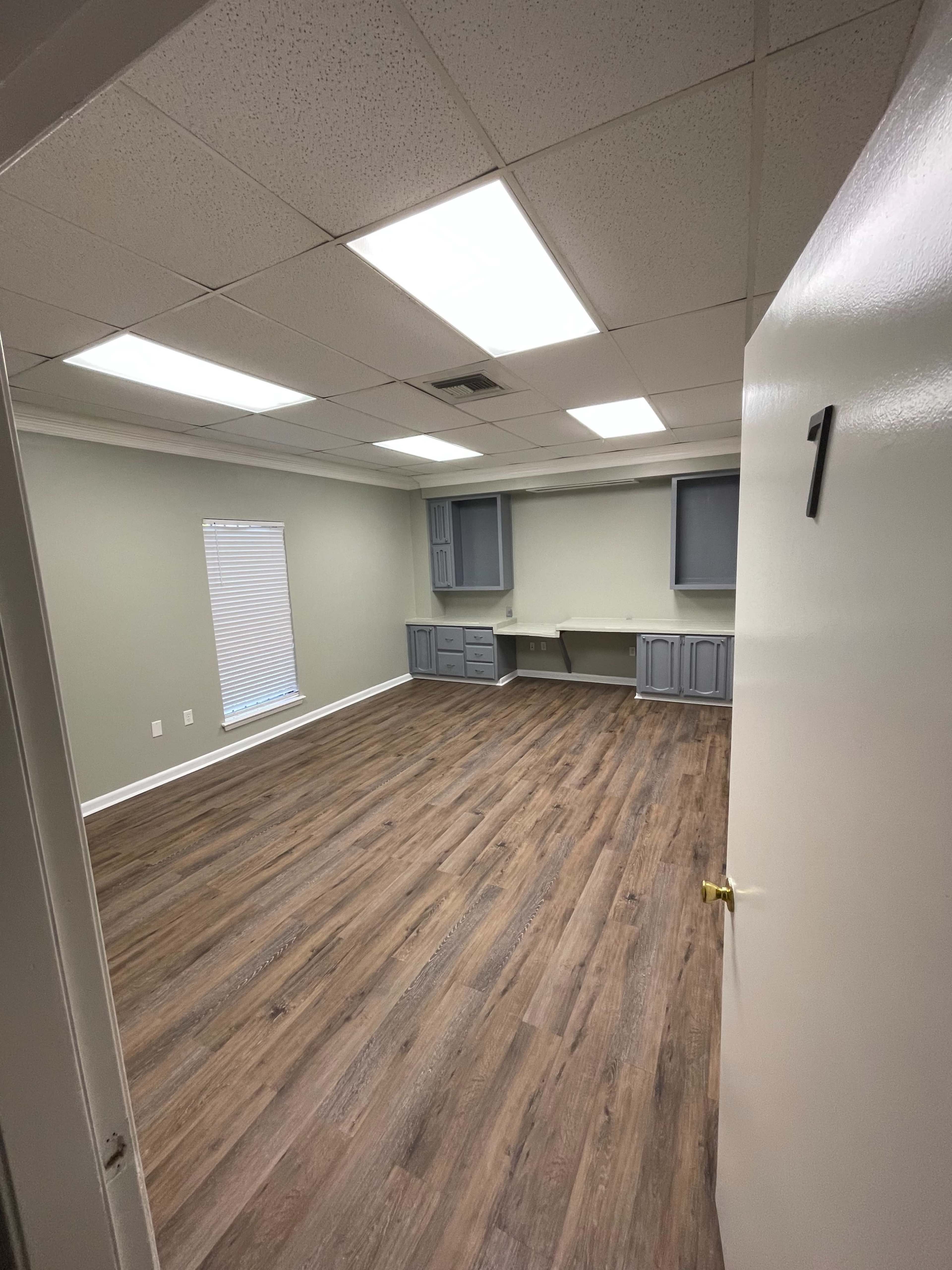 A vacant room with wooden flooring, light gray walls, and a built-in desk area is shown, illuminated by overhead fluorescent lights.