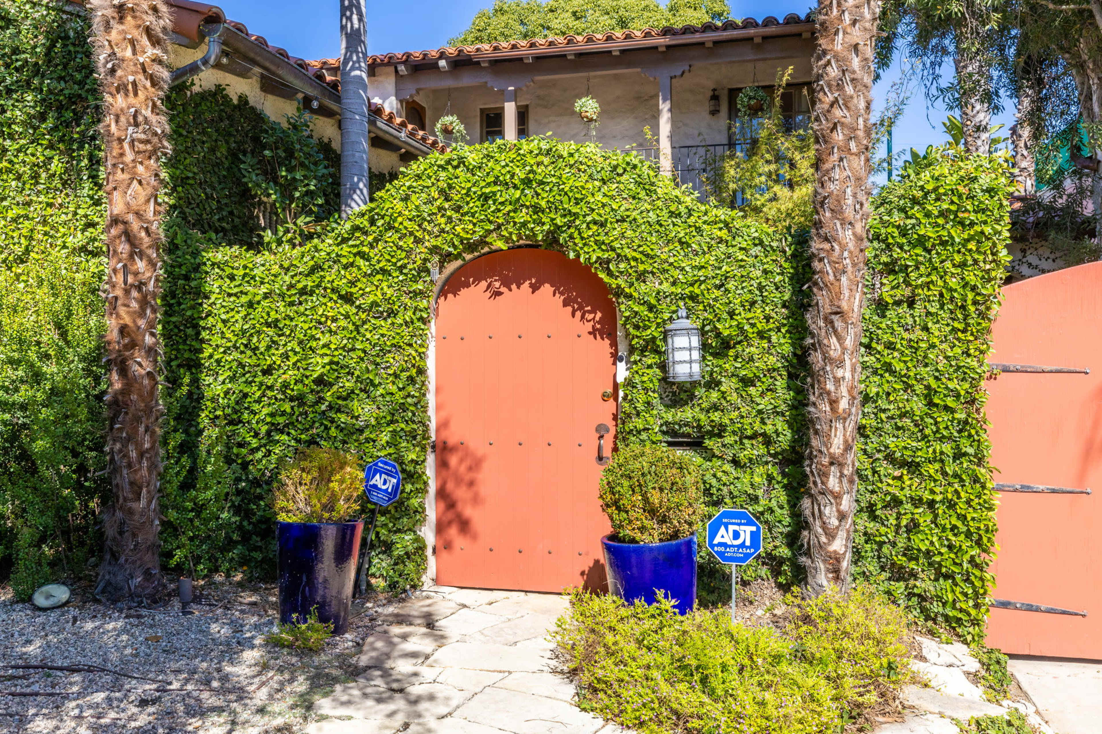 A bright orange door is set within an archway covered in green vines, flanked by tall blue planters and security signs.