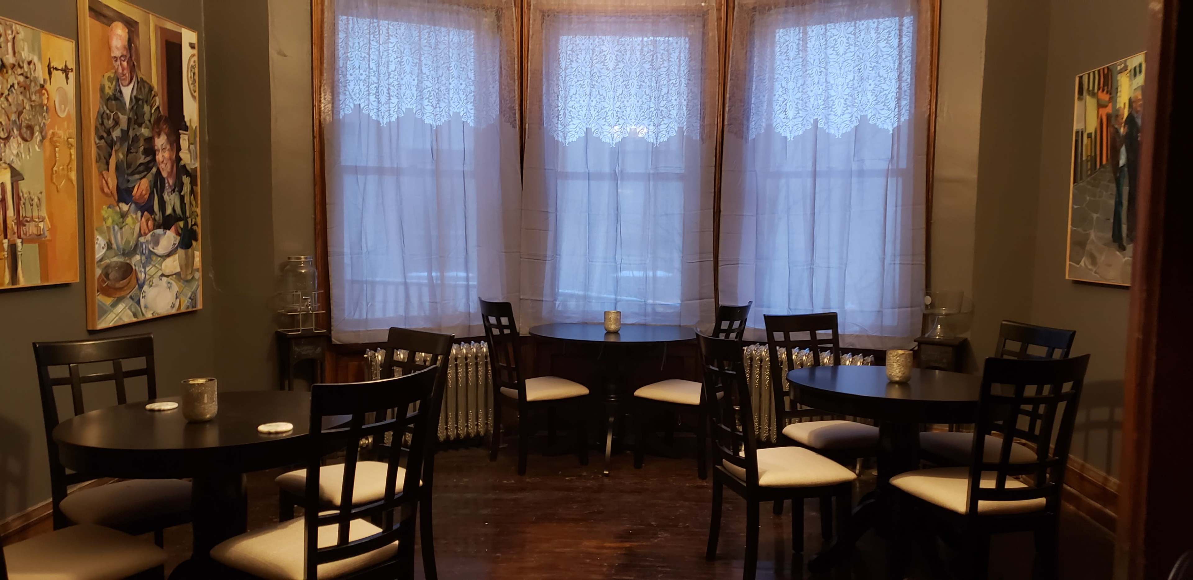 A cozy dining area with several round tables and chairs arranged around three large windows covered with sheer curtains.