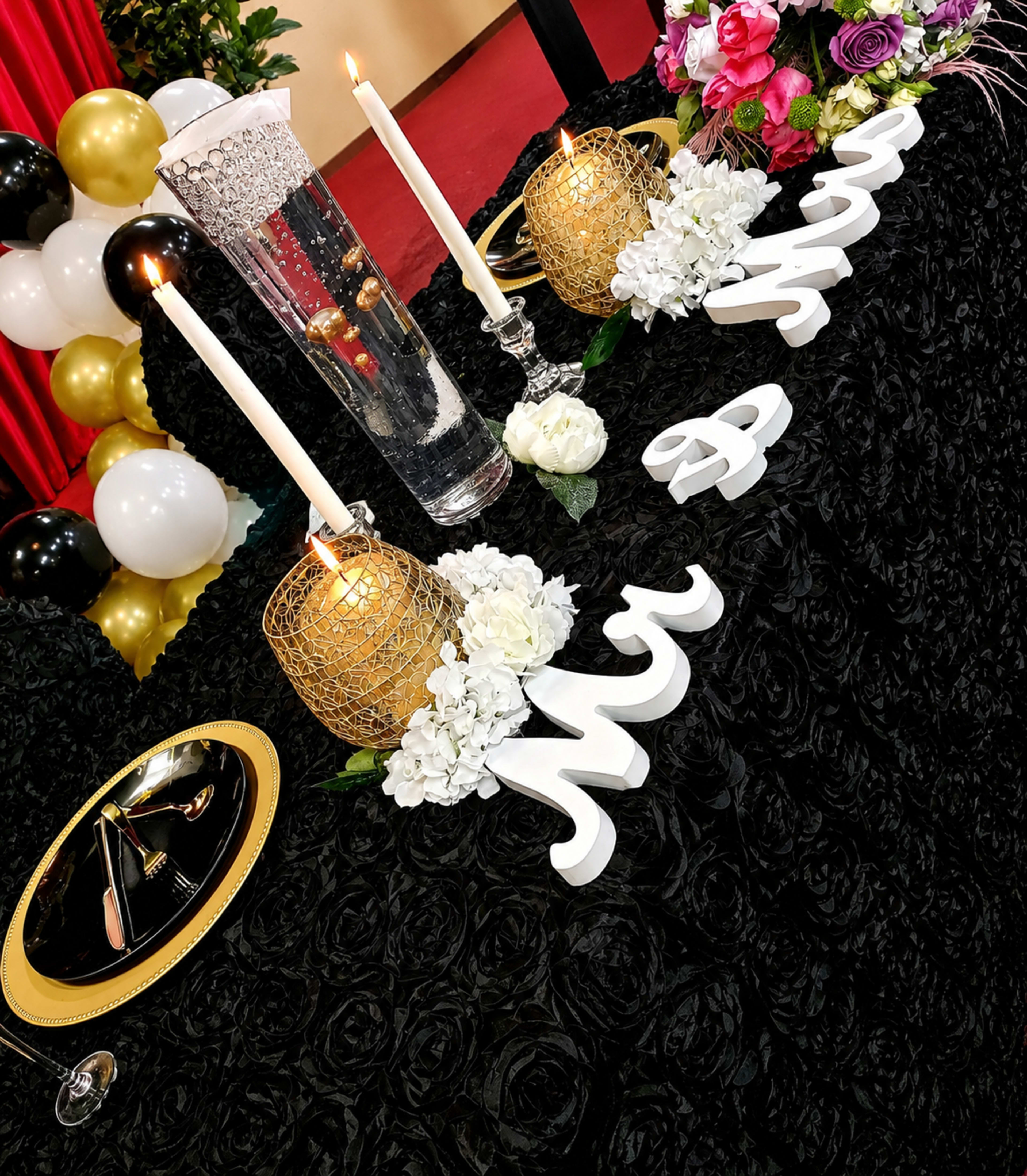 The image shows a decorated table for an event, featuring gold and white candles, floral arrangements, and decorative letters spelling "Mr & Mrs." against a black textured tablecloth.
