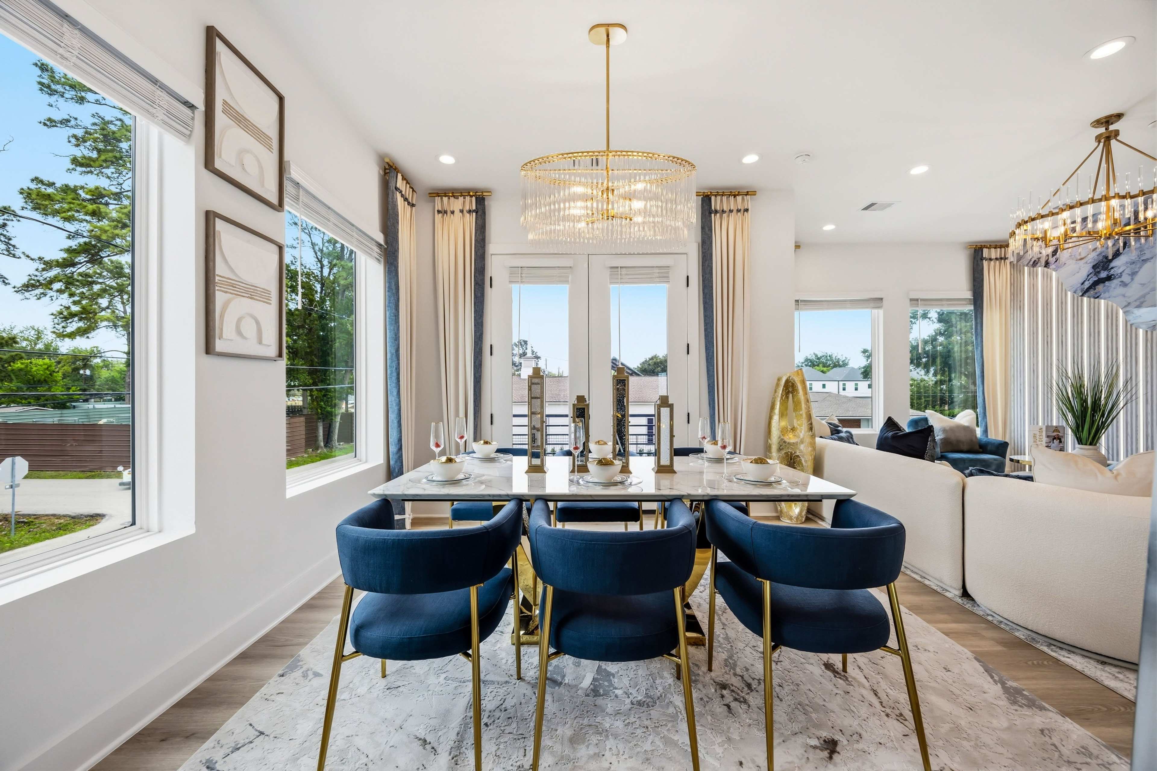 A modern dining area features a glass table surrounded by blue upholstered chairs, with a large chandelier above and natural light streaming in through the windows.