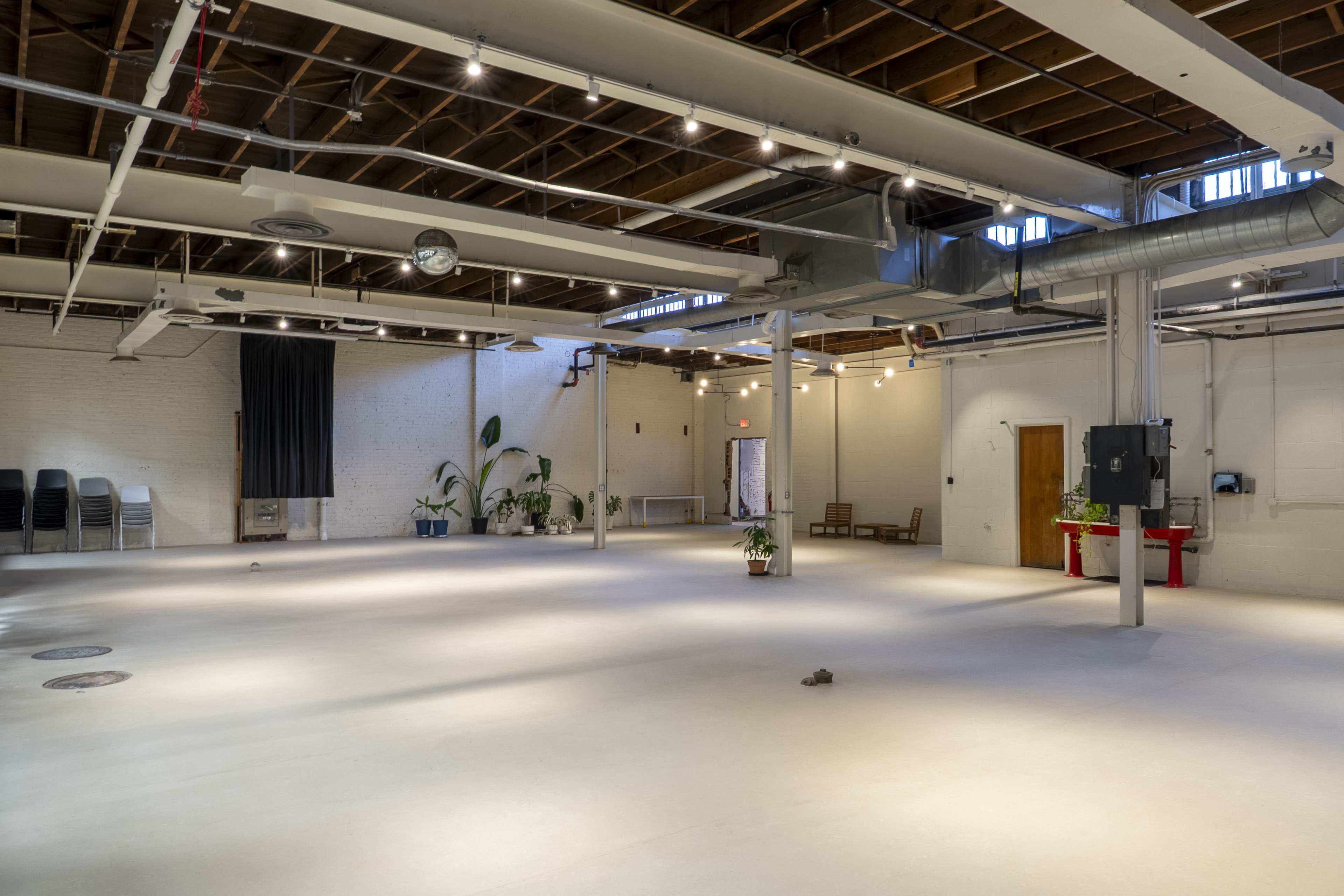 The image shows a spacious, empty room with concrete flooring, exposed beams, and minimal furnishings, featuring a few potted plants and stacks of chairs against the walls.