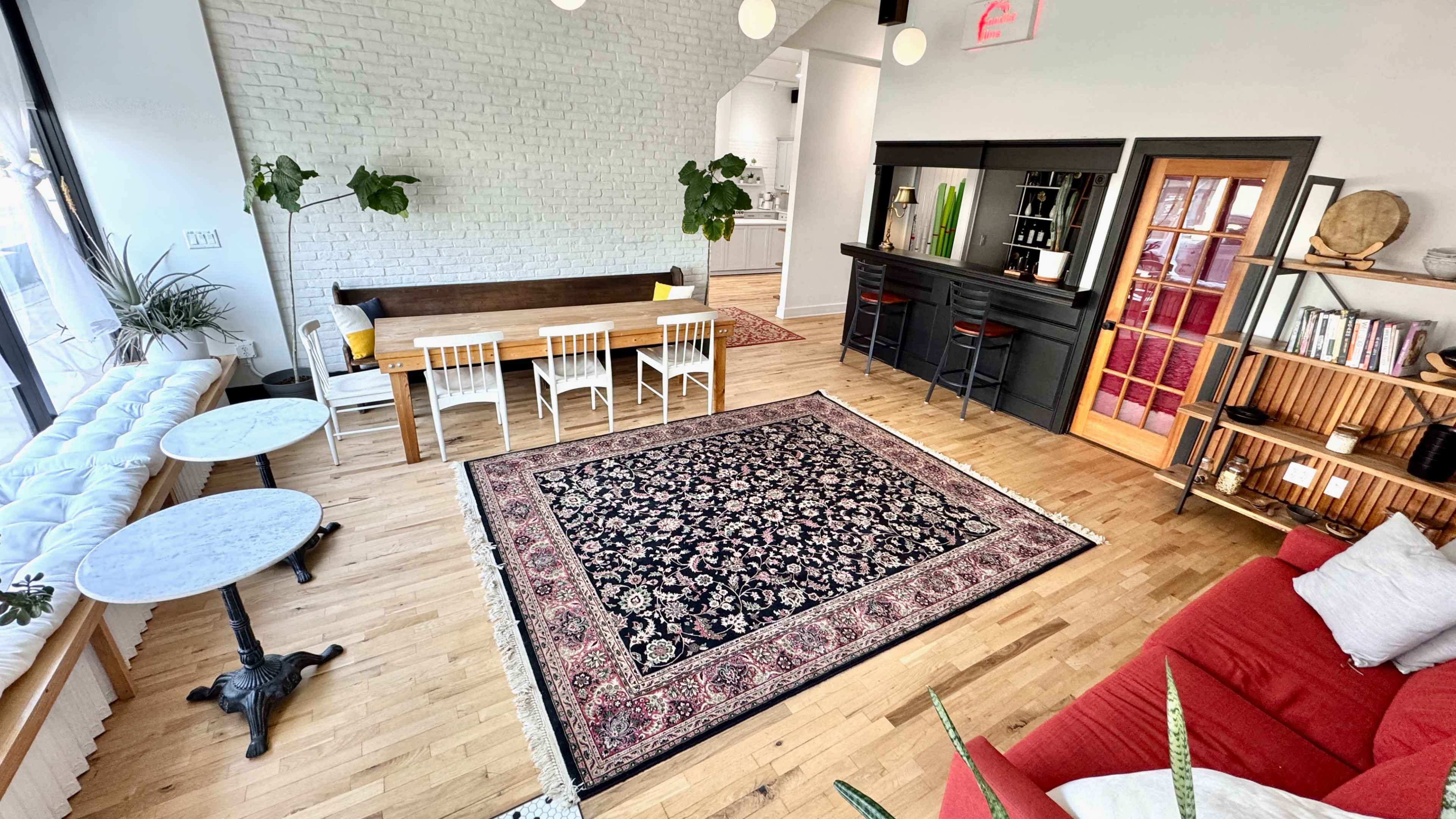 A spacious, modern living area features a hardwood floor, a large area rug, a dining table with chairs, two small tables, and a black bar area leading to another room.