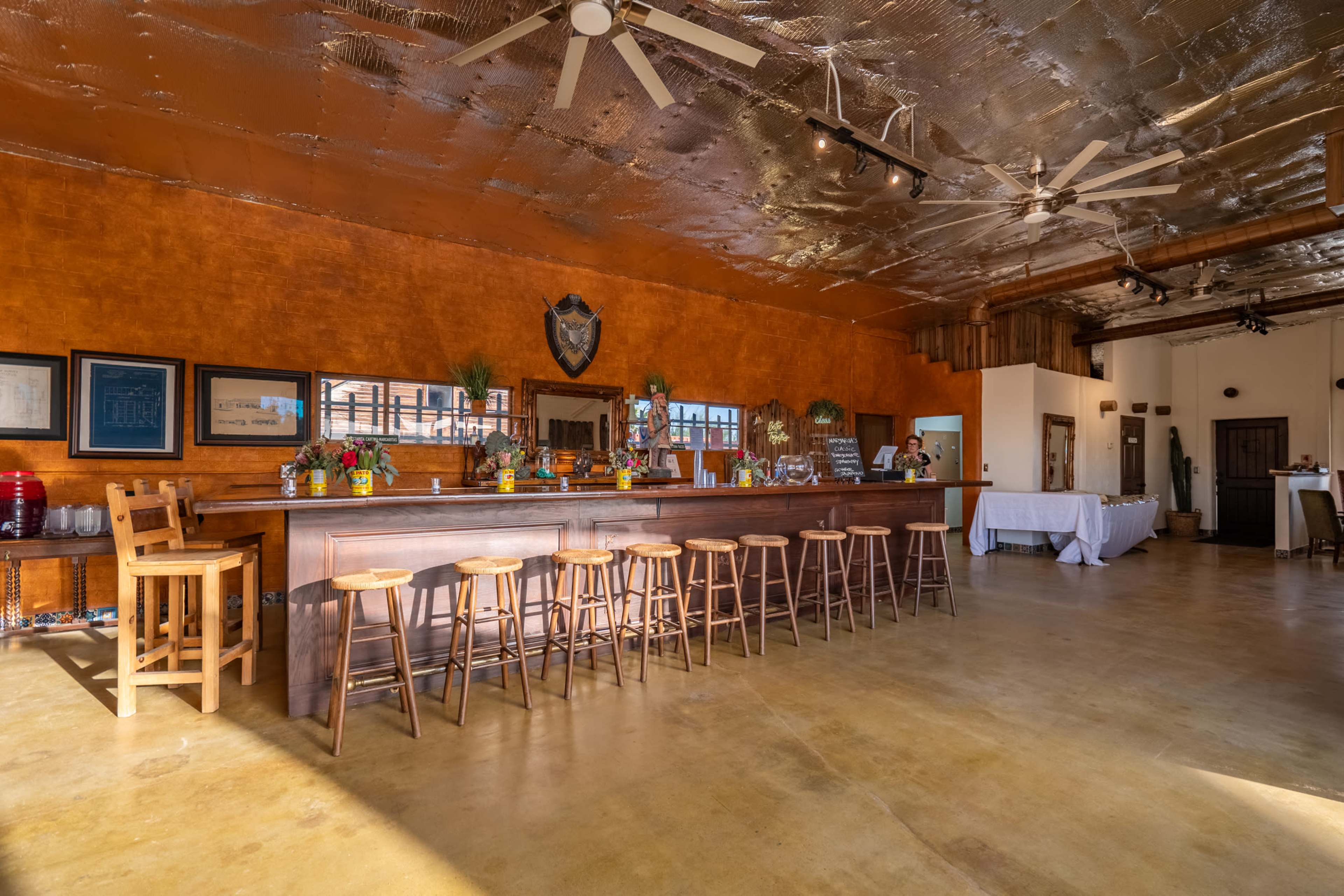 The image shows a spacious bar area with wooden stools, a countertop adorned with drinks and decorations, and a warm-colored wall.