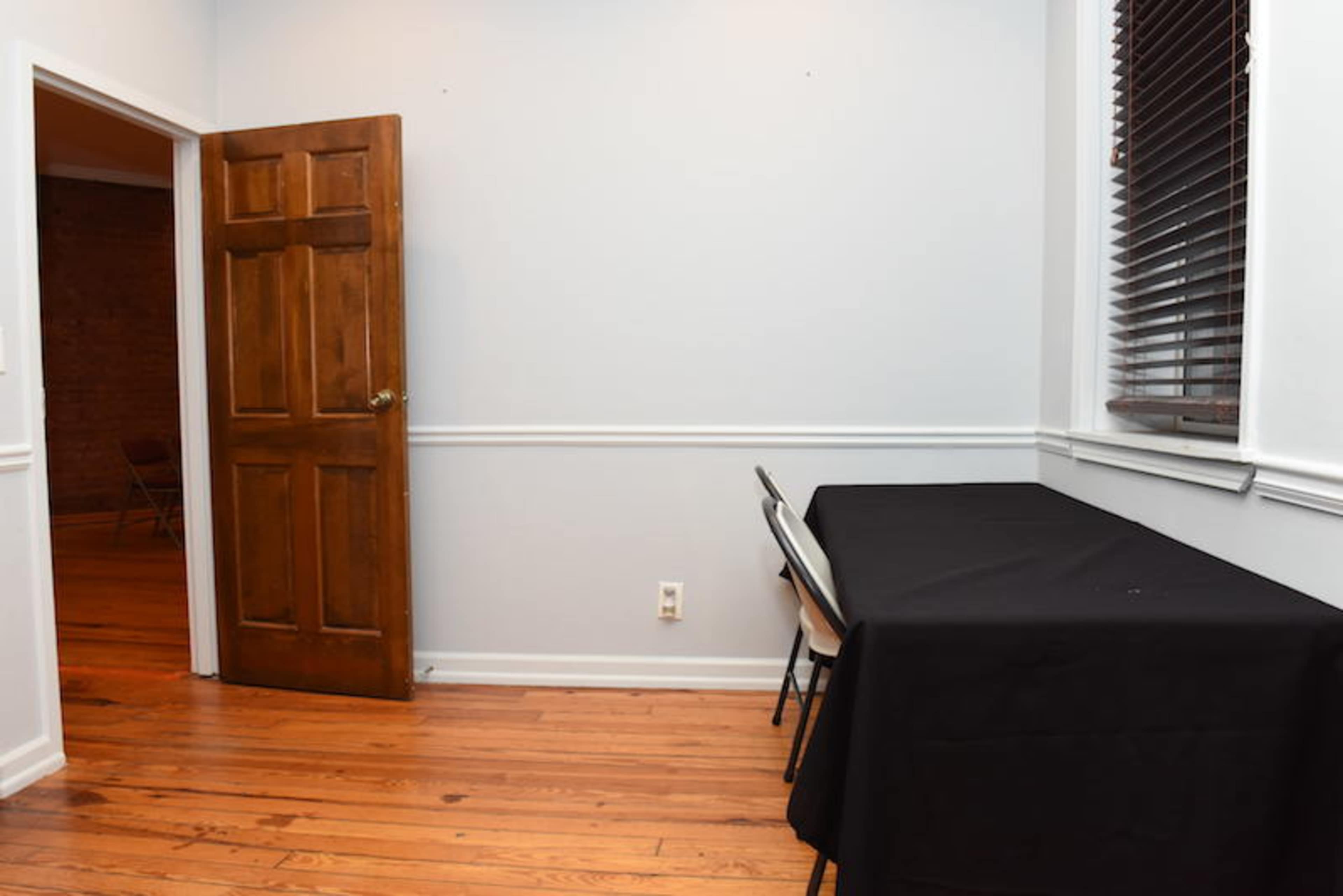 A room with a wooden door, a black tablecloth covering a table, and a window with blinds, all set against light-colored walls and wooden flooring.