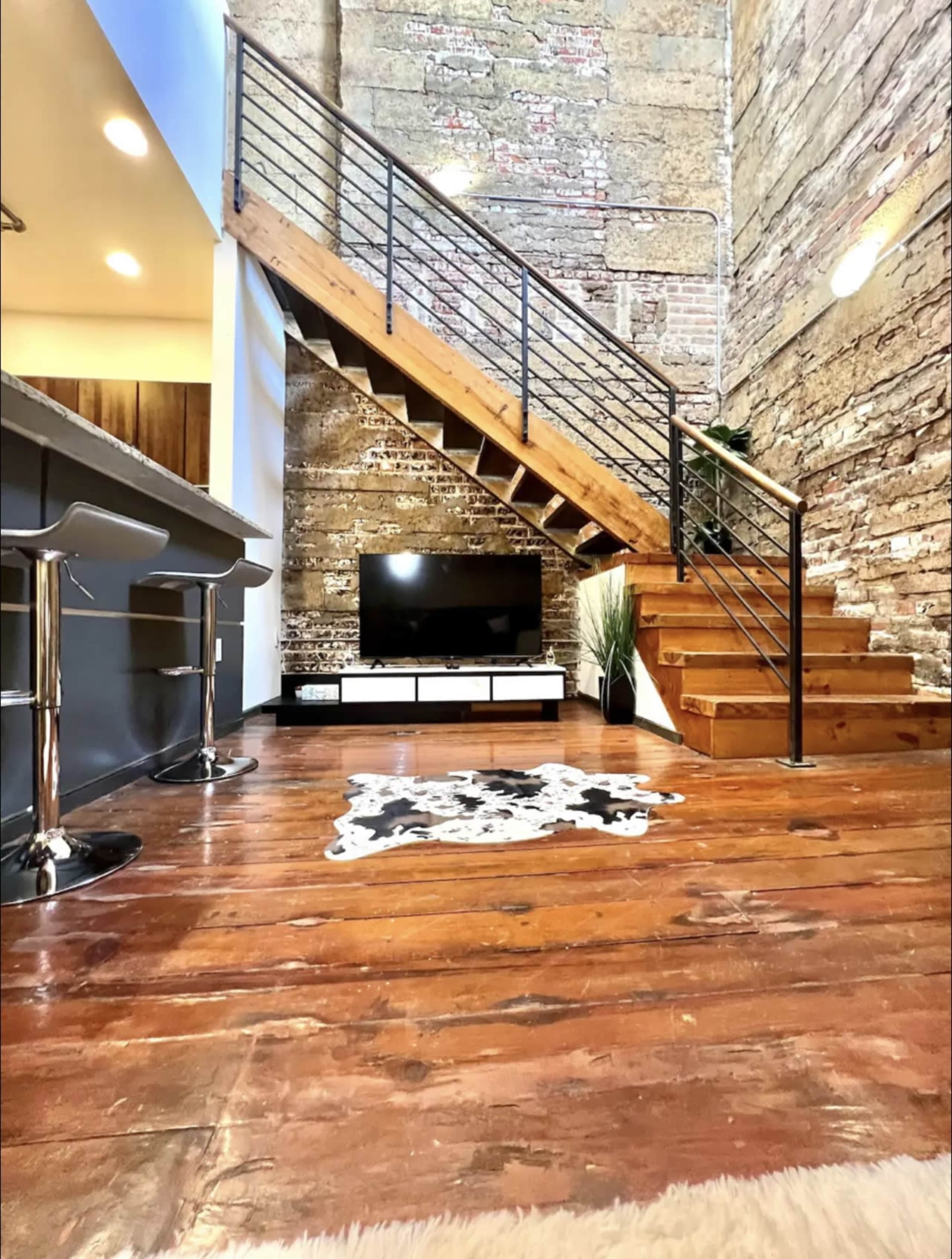 A modern interior with a staircase made of wood, a black TV mounted on a white cabinet, and a cowhide rug on a wooden floor, set against exposed brick walls.