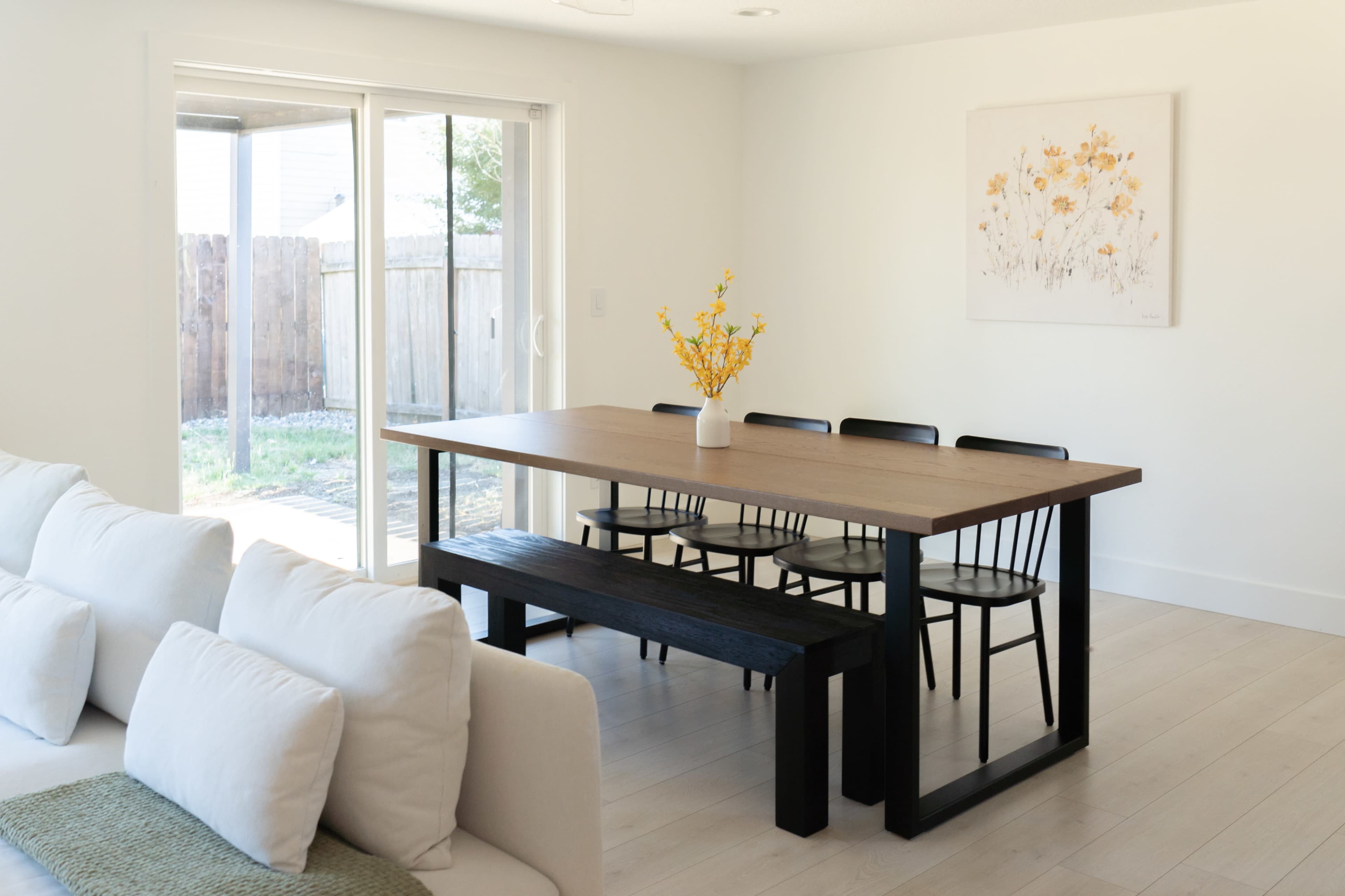 A modern dining area features a wooden table with six black chairs and a bench, complemented by a vase of yellow flowers, against a backdrop of sliding glass doors leading to an outdoor space.