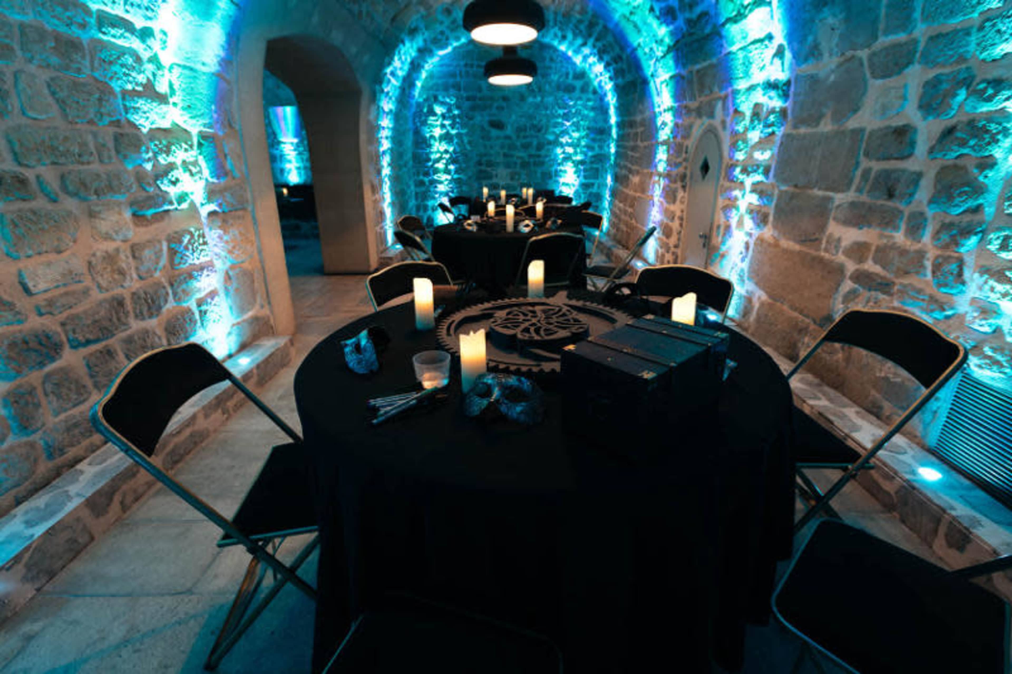 A dimly lit room with stone walls, featuring round tables set with black tablecloths, illuminated by blue lights and surrounded by metal chairs.