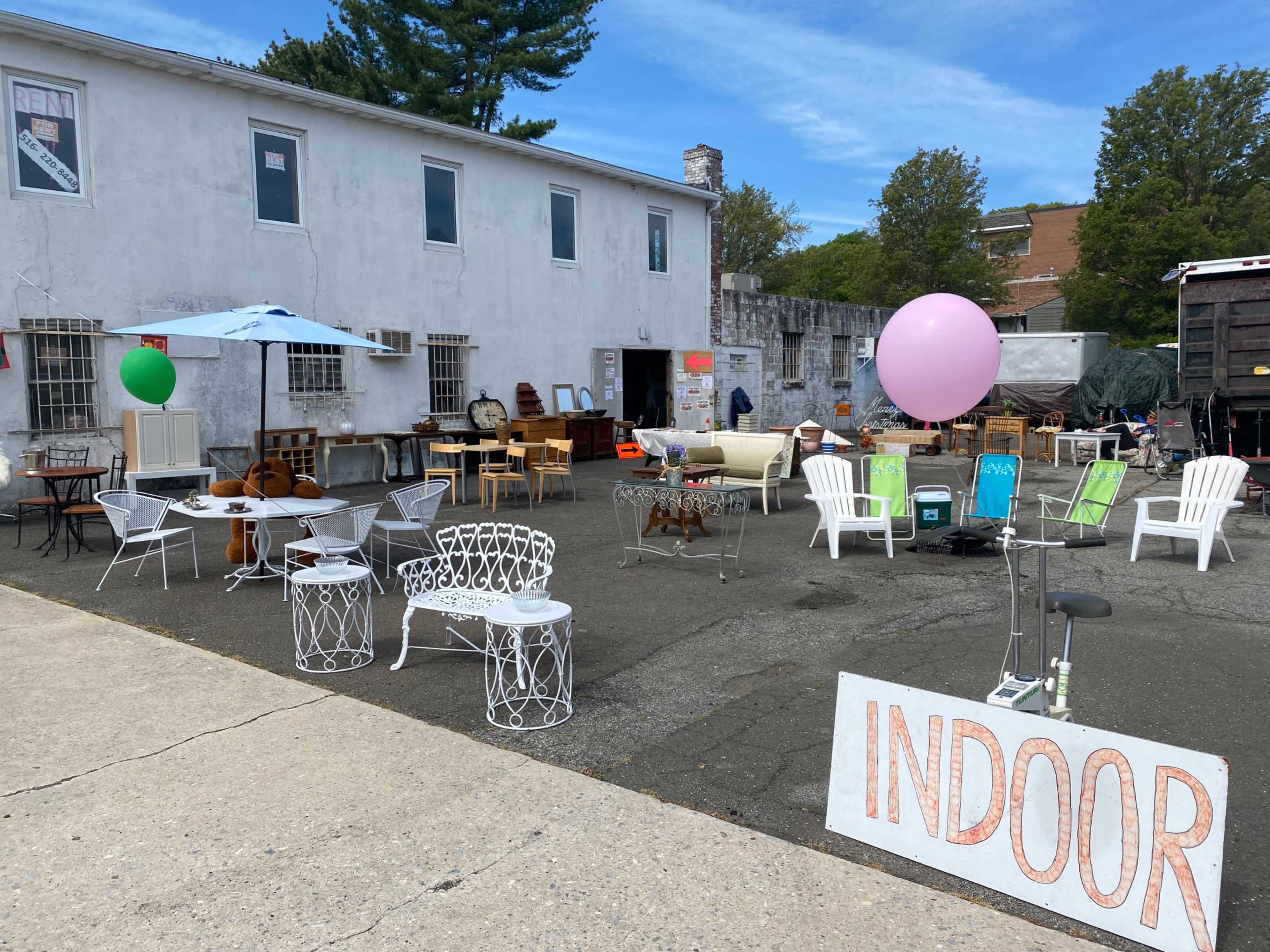 The scene shows a courtyard filled with various pieces of furniture for sale, including chairs, tables, and decorative items, with a sign indicating "INDOOR."