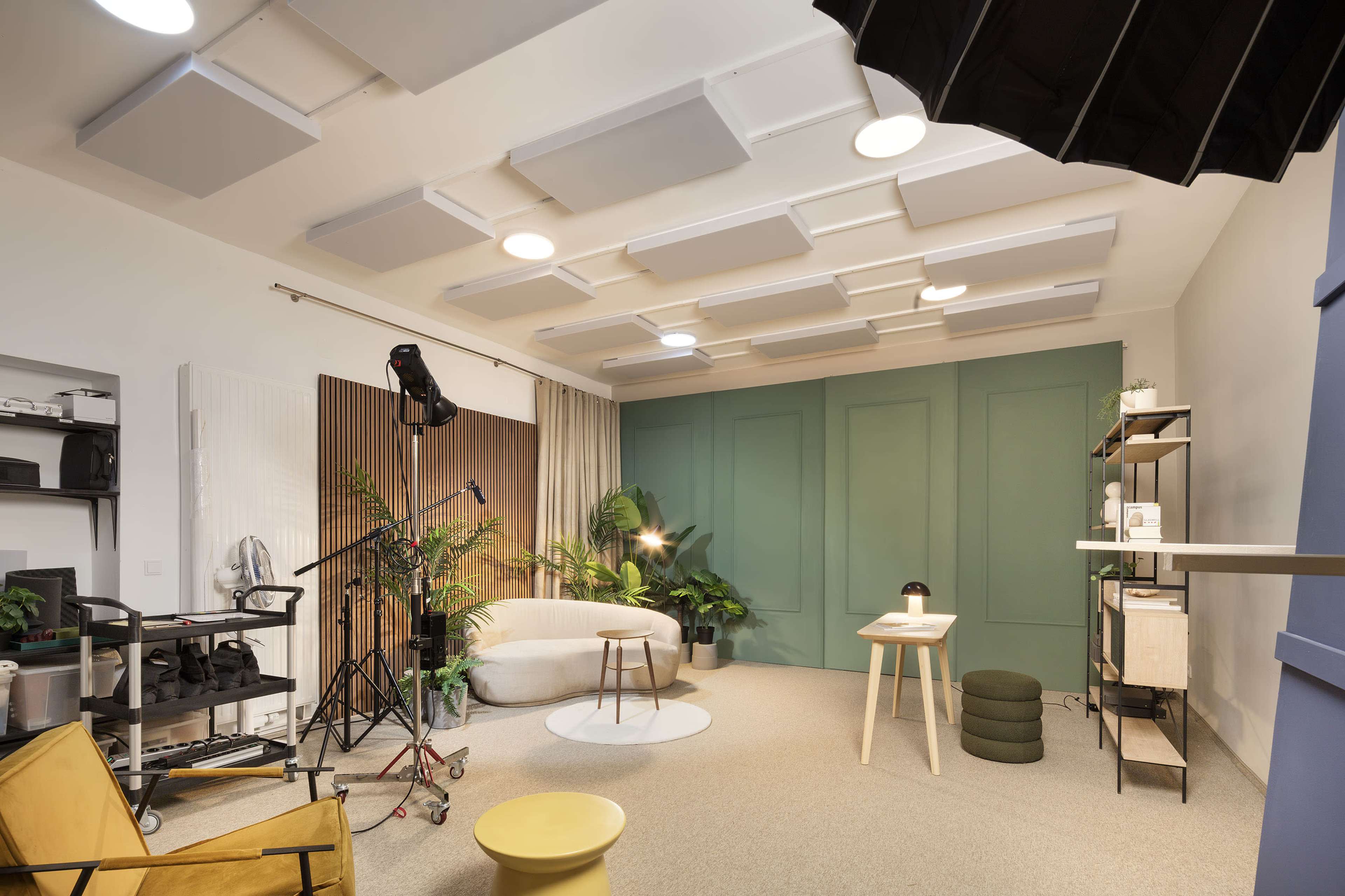 A modern studio space with a green accent wall, various plants, and a circular sofa, accompanied by a lighting setup and shelves with equipment.
