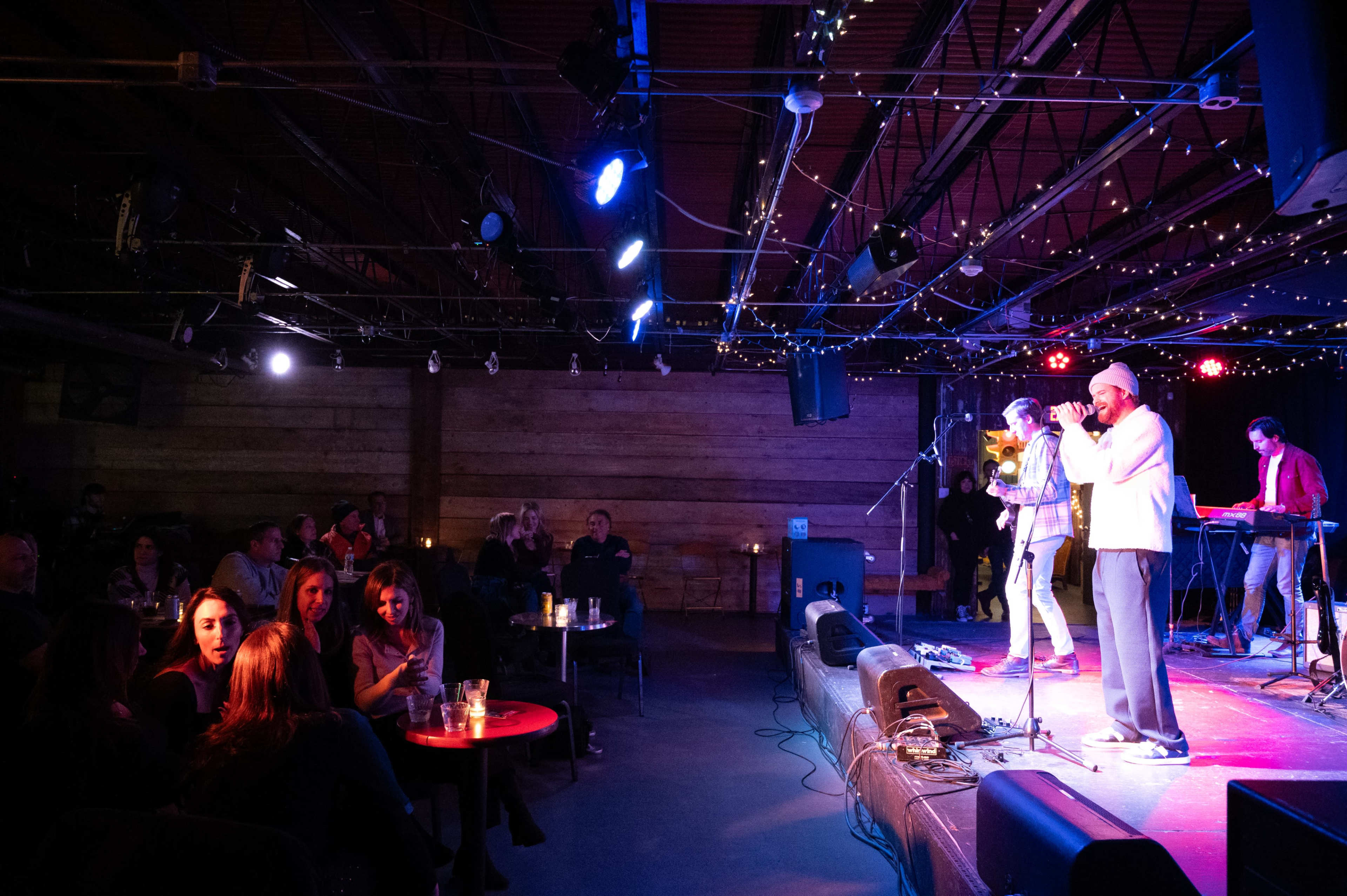 A band performs on stage in a lively venue while an audience engages at tables nearby.