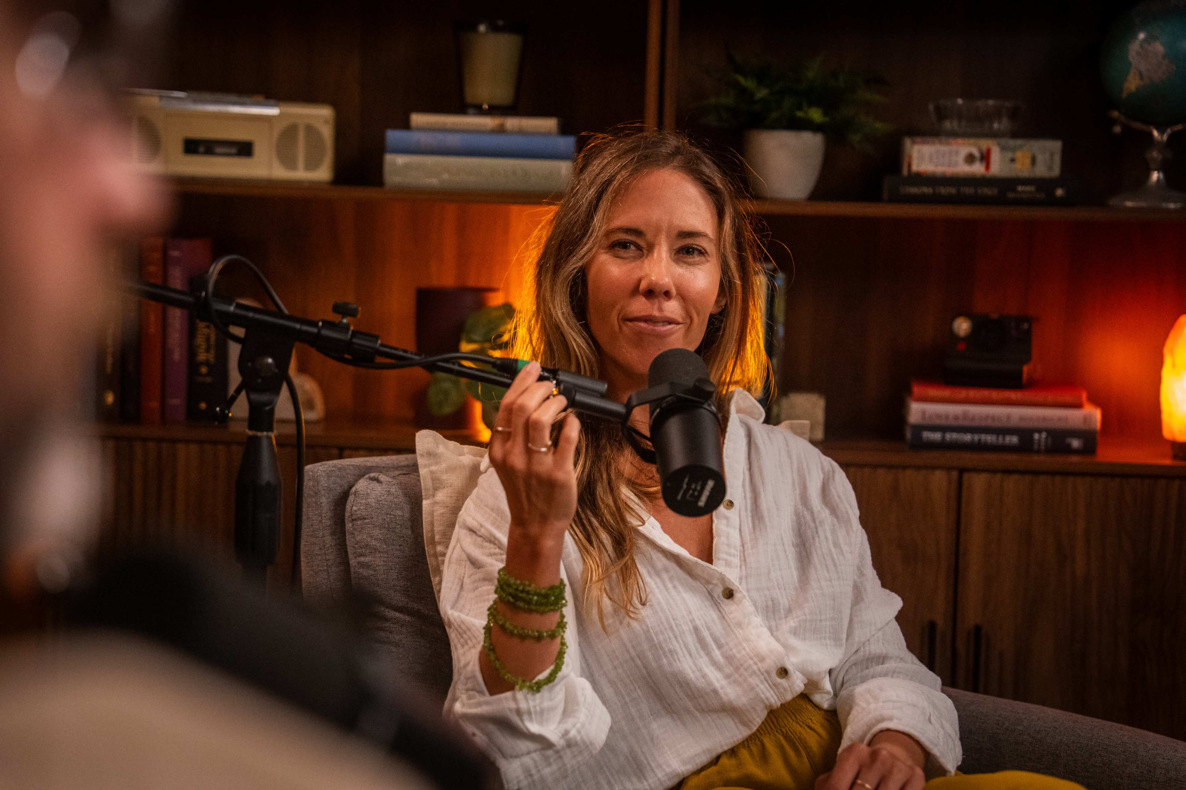 A woman with long hair sits on a couch, speaking into a microphone in a well-lit room with bookshelves and plants in the background.
