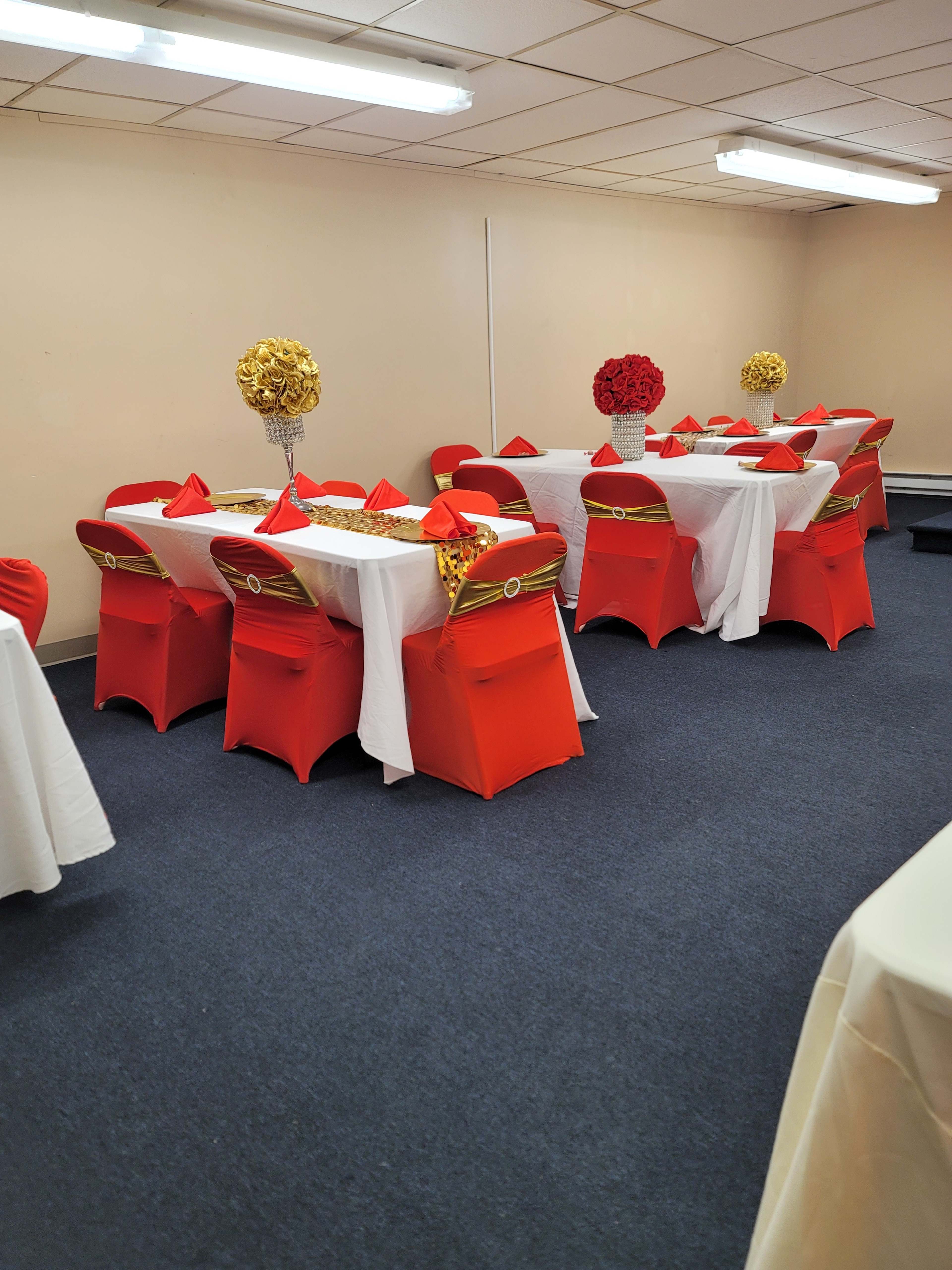 The room features several tables covered with white tablecloths, surrounded by red and gold chairs, arranged for an event celebration.