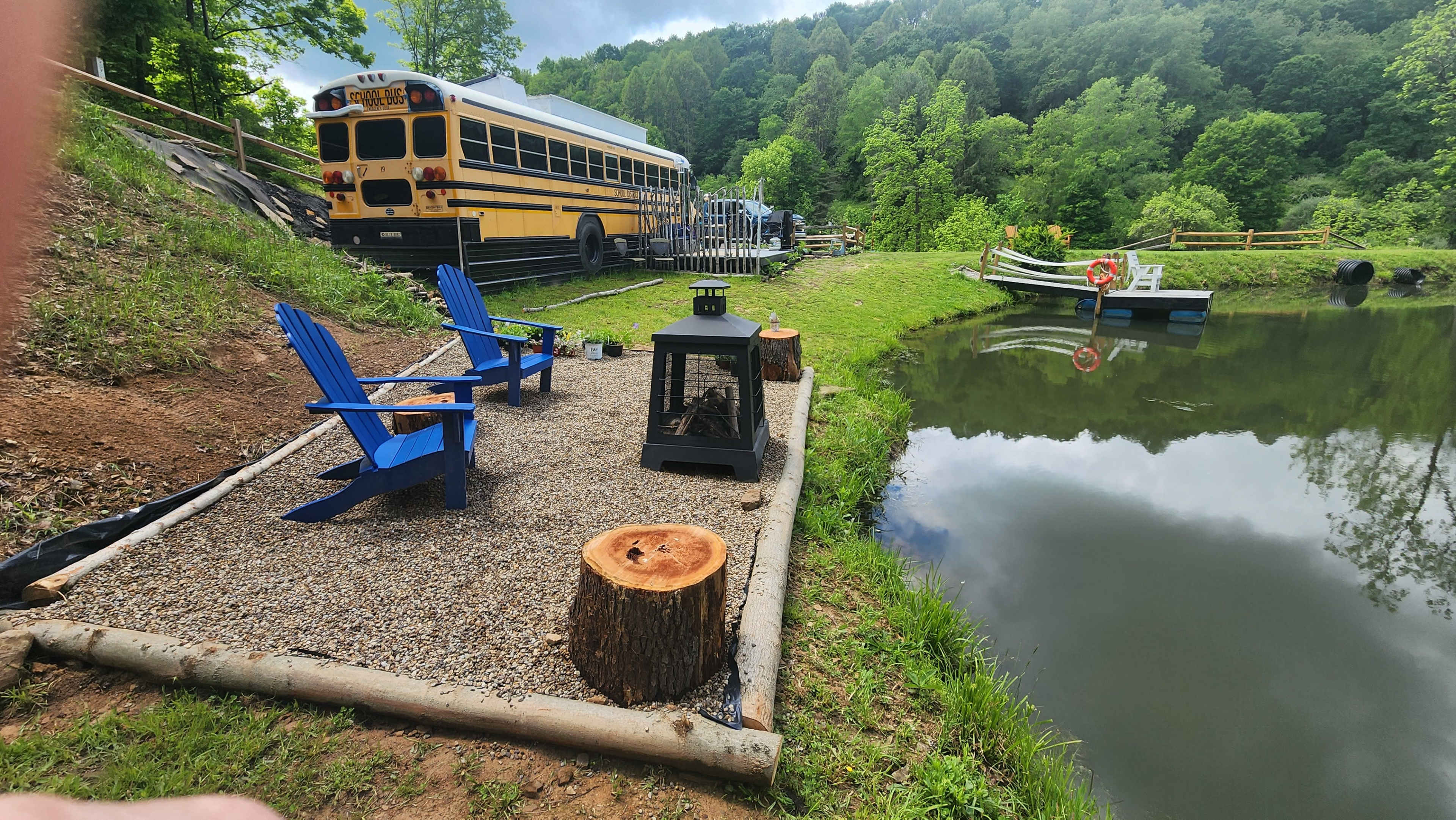 The scene features two blue Adirondack chairs and a small fire pit set on gravel by a pond, with a yellow school bus parked nearby and a wooden dock in the background.