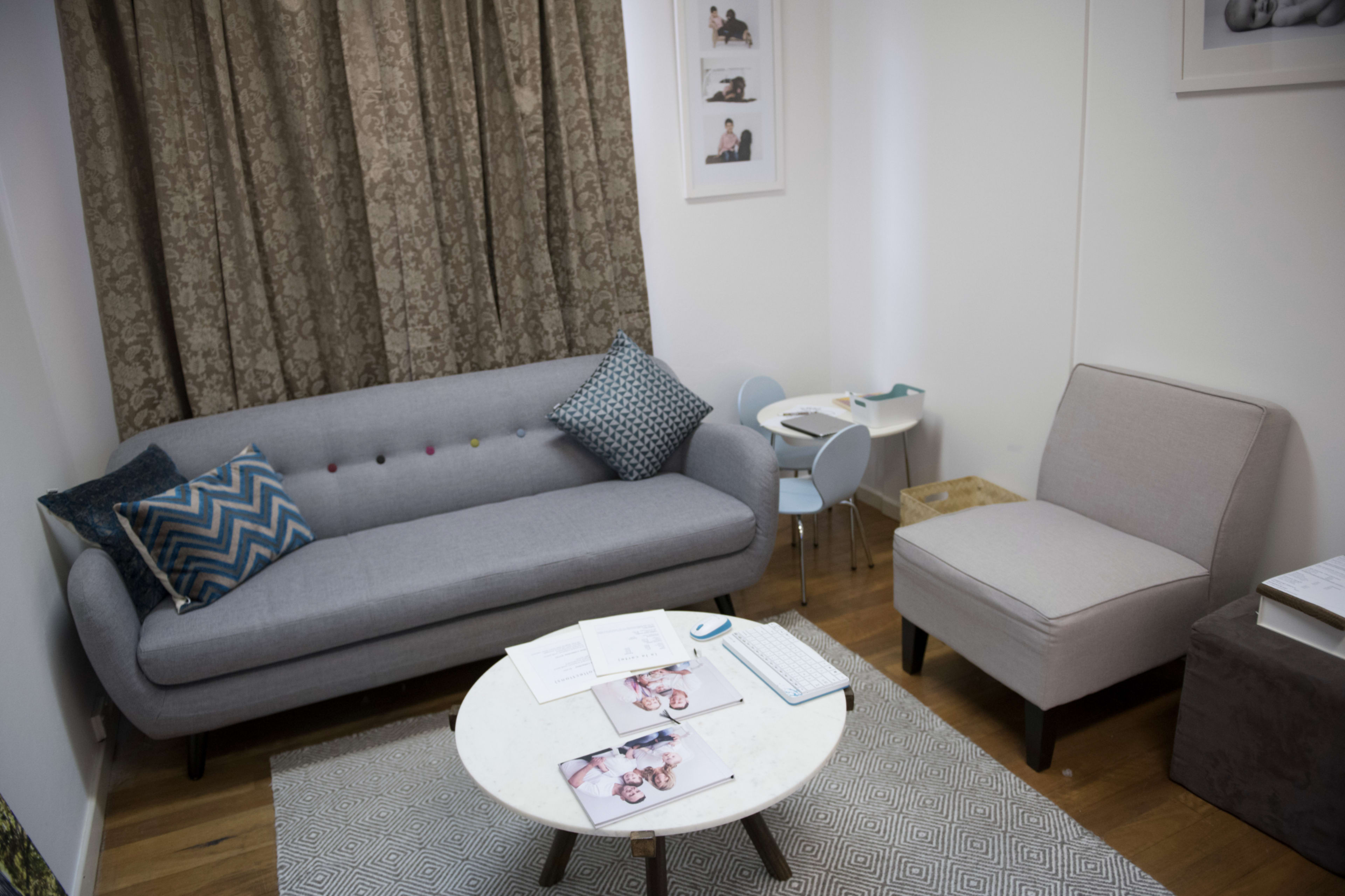 The image shows a living room with a gray sofa, a light gray armchair, a round coffee table, and a small child’s table with chairs, all set against a backdrop of patterned curtains.