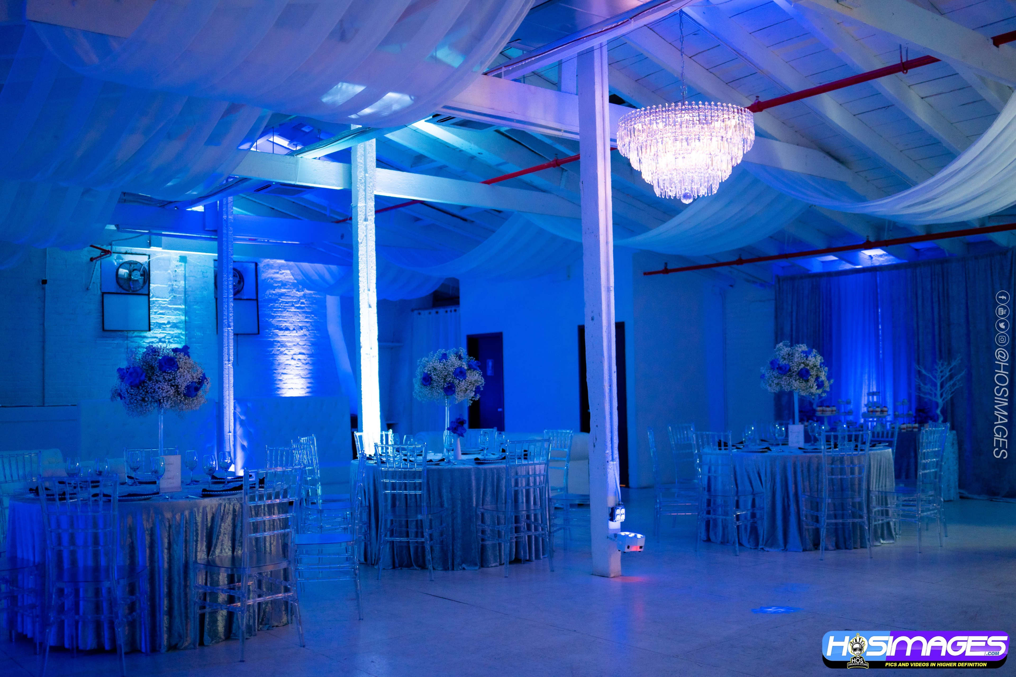 A banquet hall featuring circular tables with silver tablecloths, clear chairs, and a chandelier, all illuminated by blue lighting.