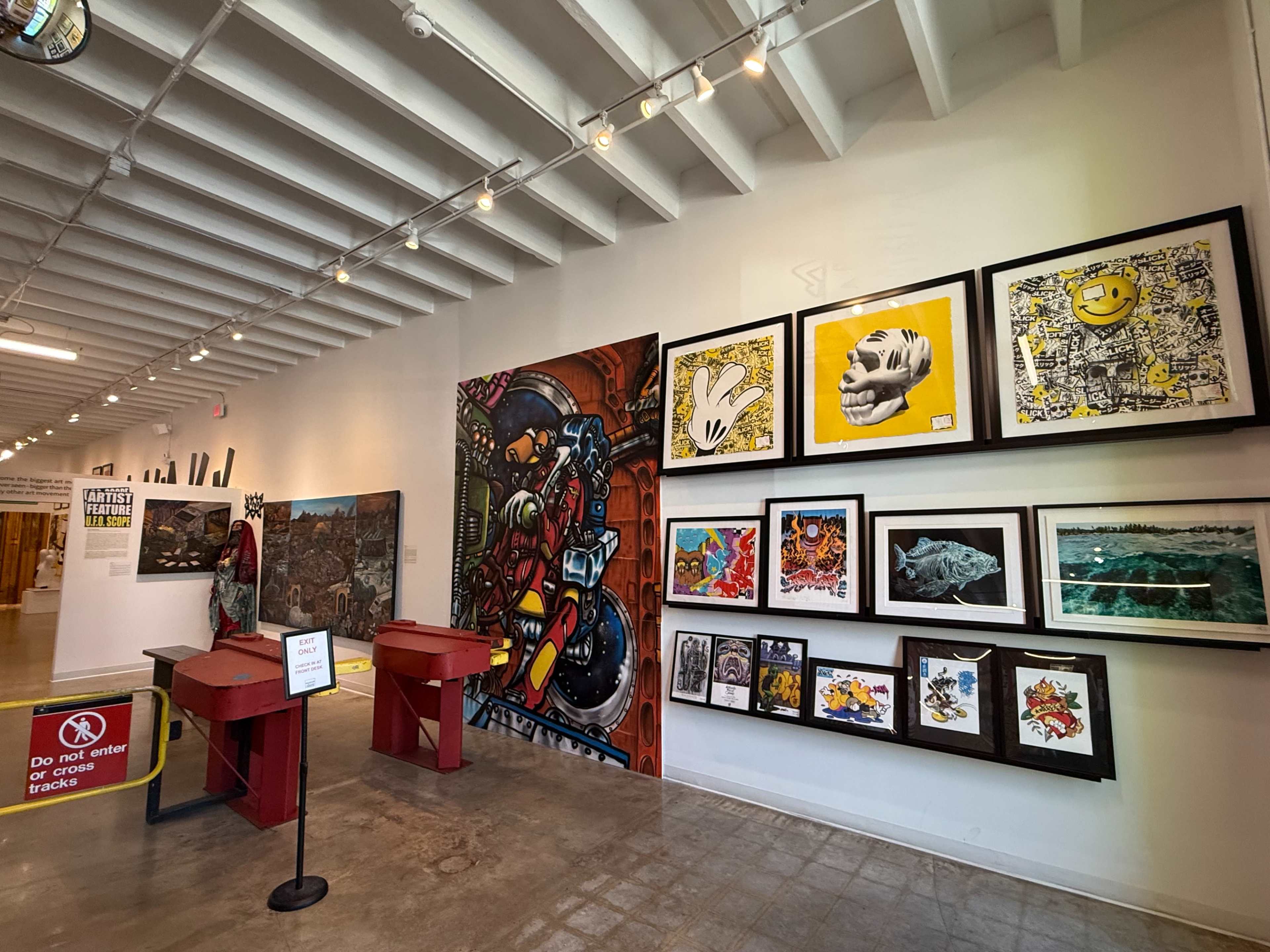 The image shows an interior gallery with a wall displaying a variety of framed artworks, including paintings and prints, alongside two red display stands at the foreground.