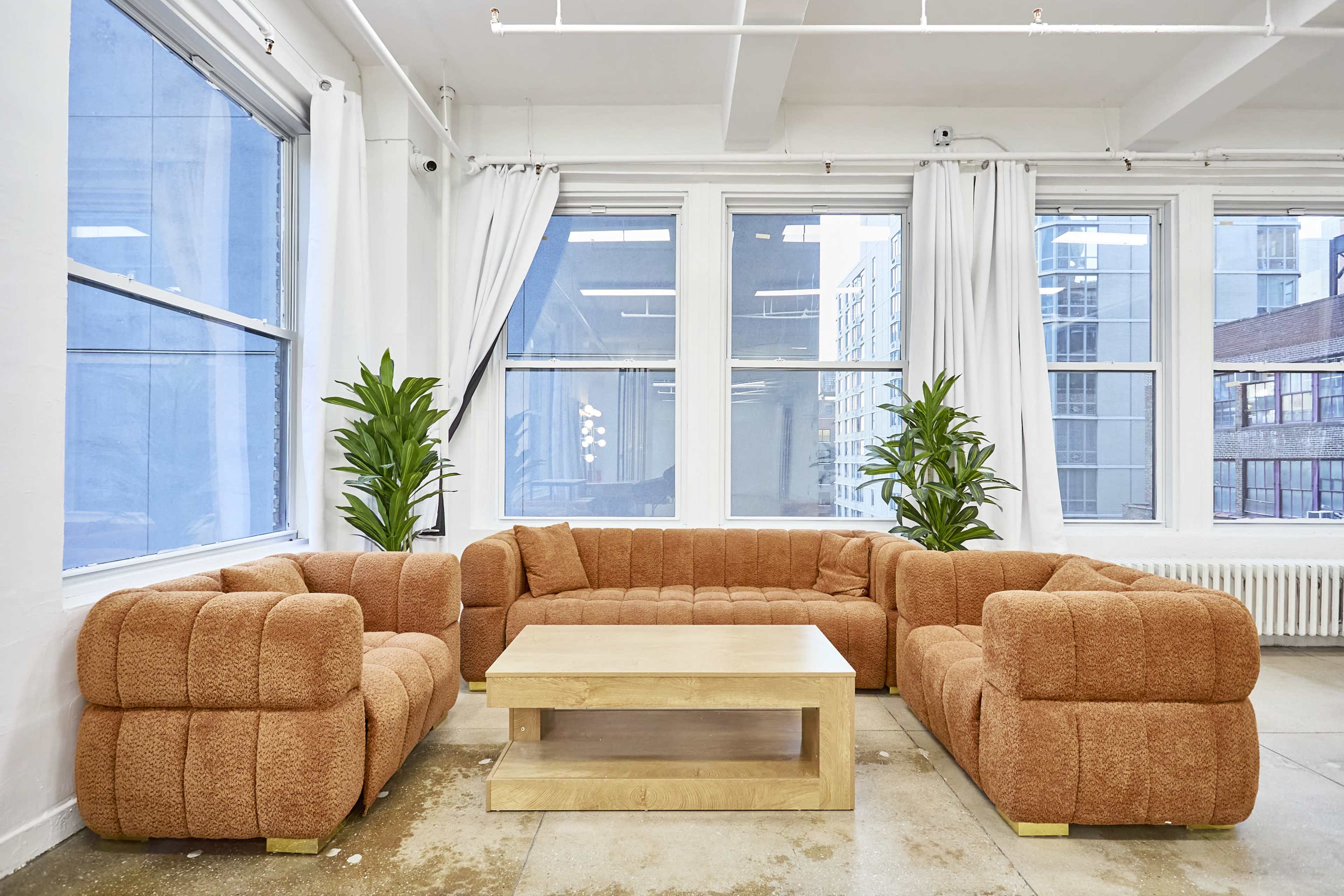 A spacious room features a large, orange sectional sofa arranged around a wooden coffee table, with plants positioned in the corners and large windows allowing natural light to enter.