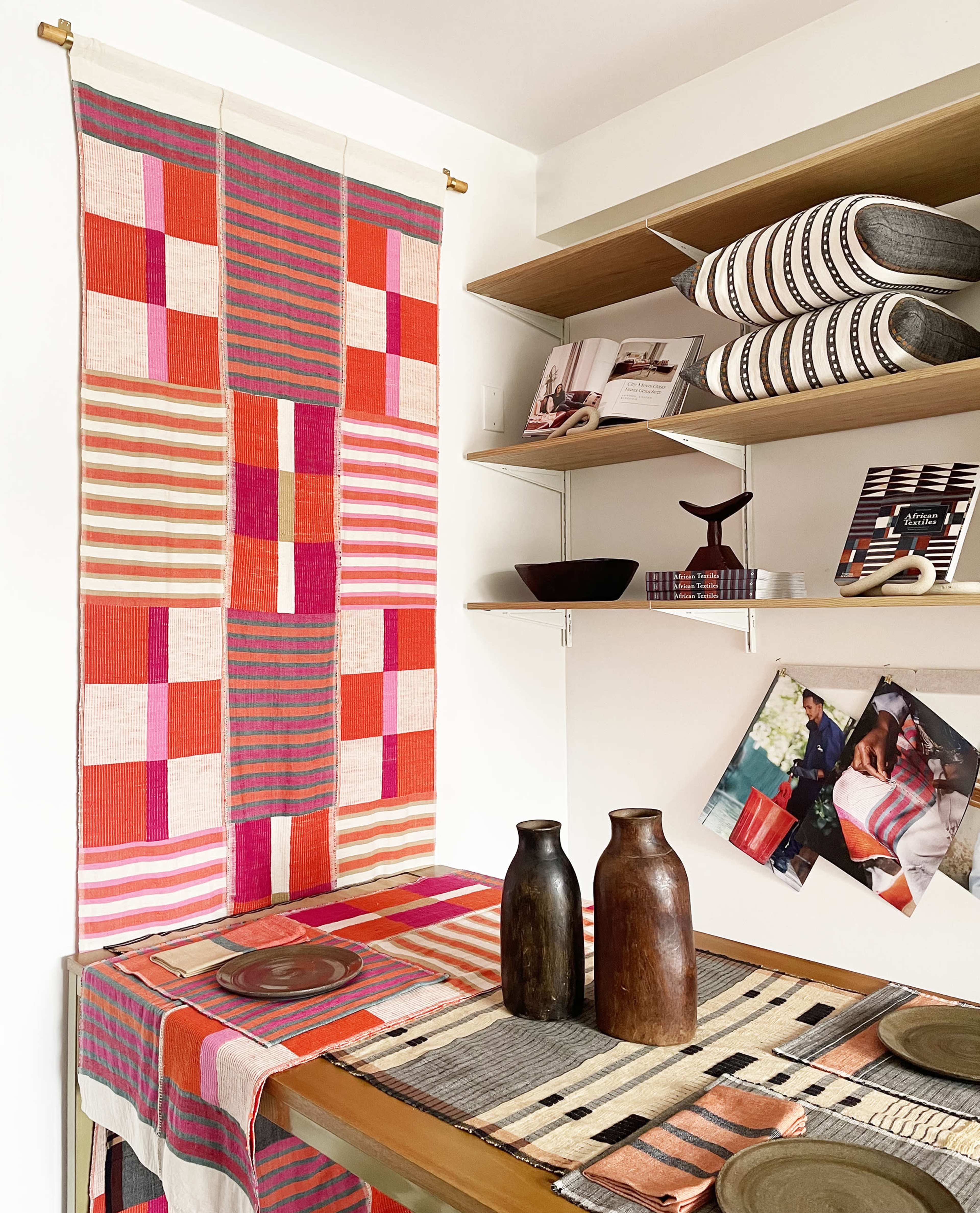 The scene features a vibrant textile hanging on the wall beside a wooden table set with plates and two decorative vases, accompanied by shelves displaying patterned bowls and magazines.
