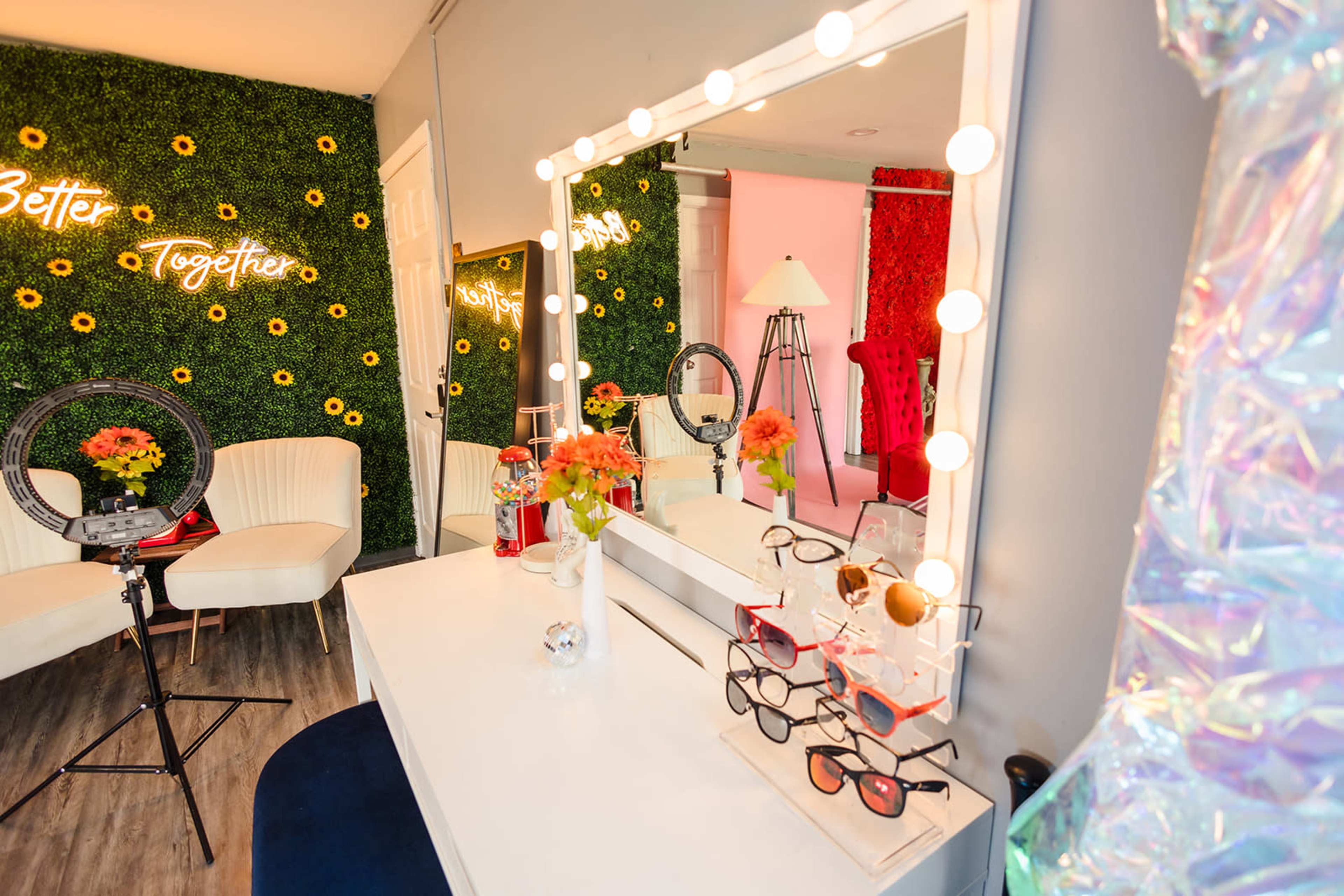 A bright vanity space with a large mirror surrounded by lights, a display of sunglasses, and vibrant flower arrangements against a backdrop of green walls with decorative text.