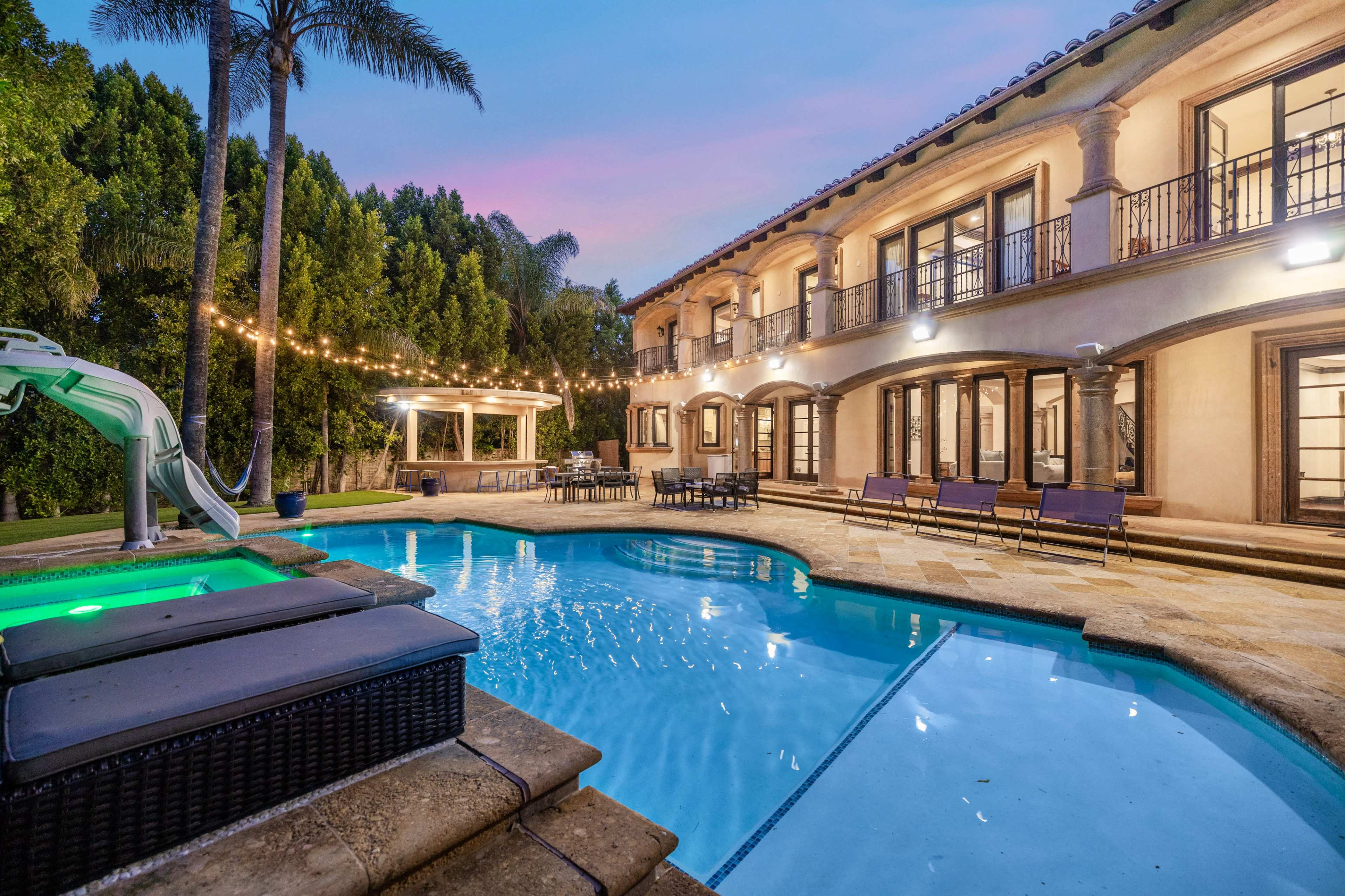A spacious backyard features a pool with a slide, lounge chairs, and a gazebo surrounded by palm trees and string lights.