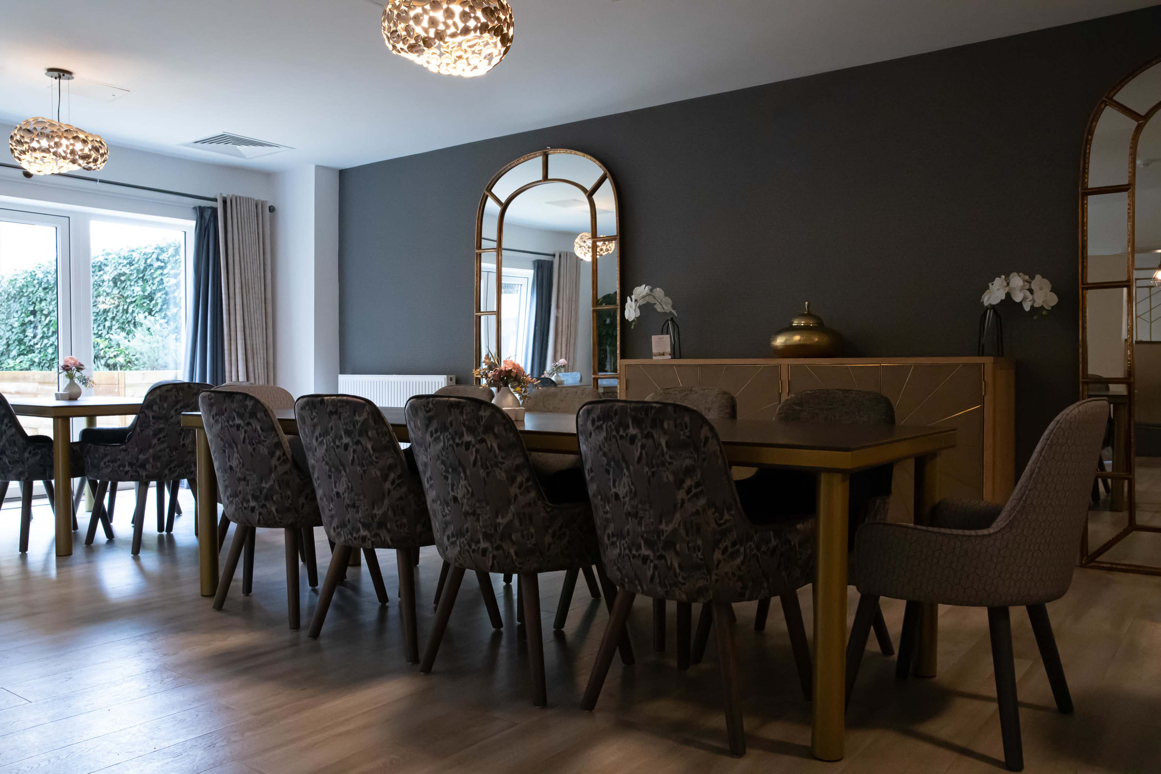 A modern dining area features a long table surrounded by upholstered chairs, with large windows and decorative mirrors on the walls.