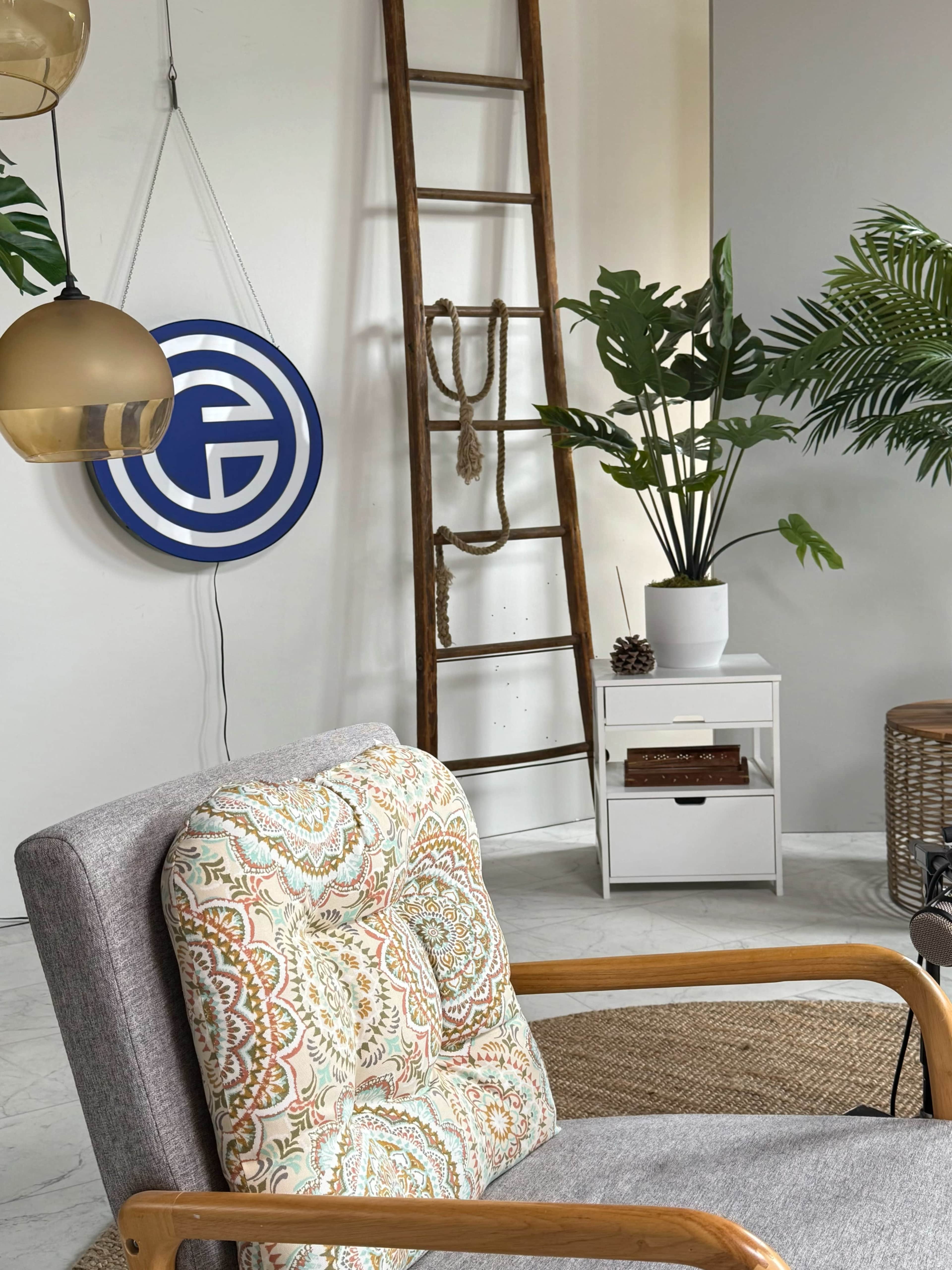 The image features a cozy living space with a gray armchair, a wooden ladder, a decorative wall sign, various houseplants, and a small white side table.