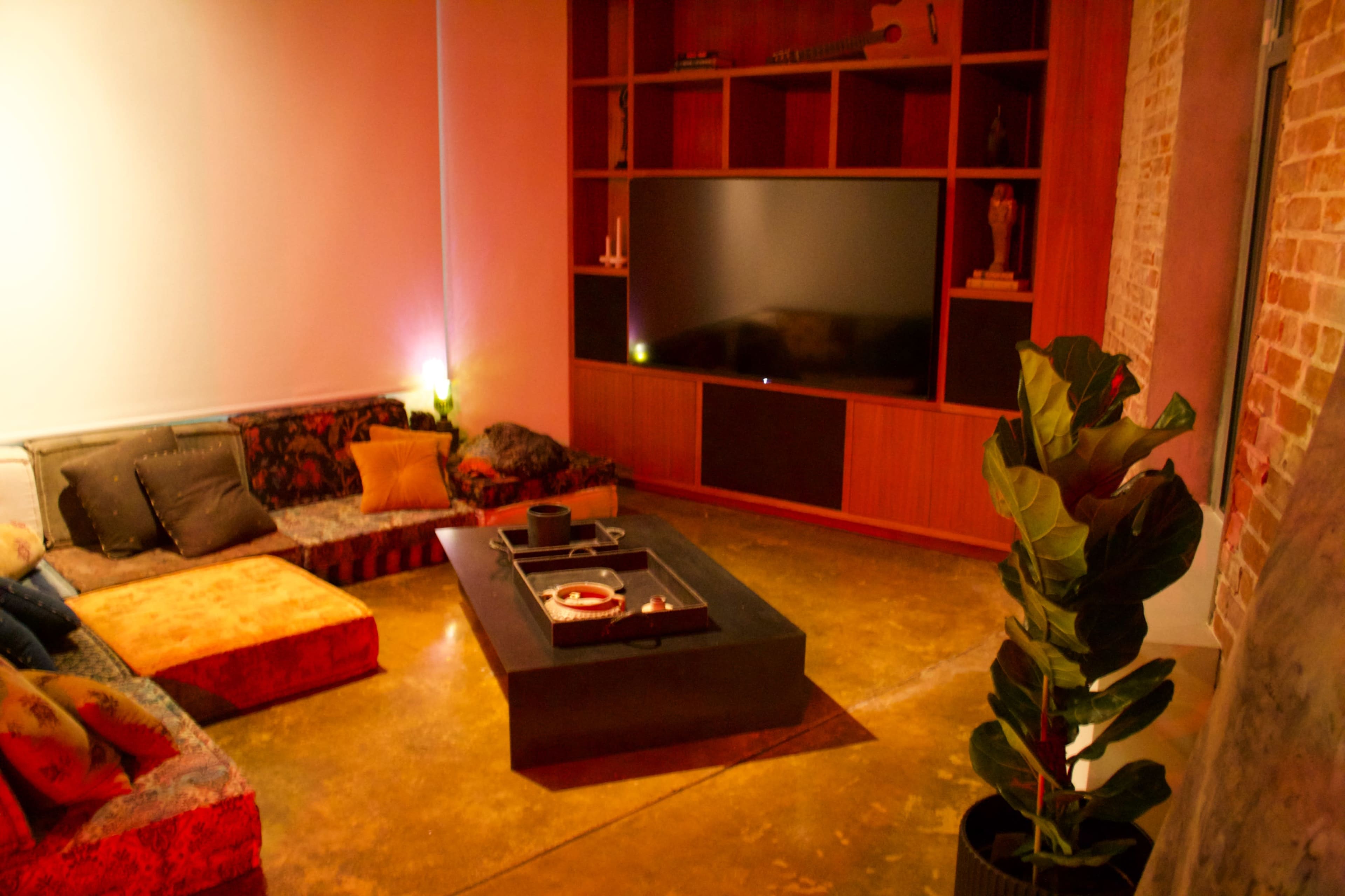 A modern living room features a large television mounted on a wooden wall unit, surrounded by a cozy seating area with colorful cushions and a coffee table.
