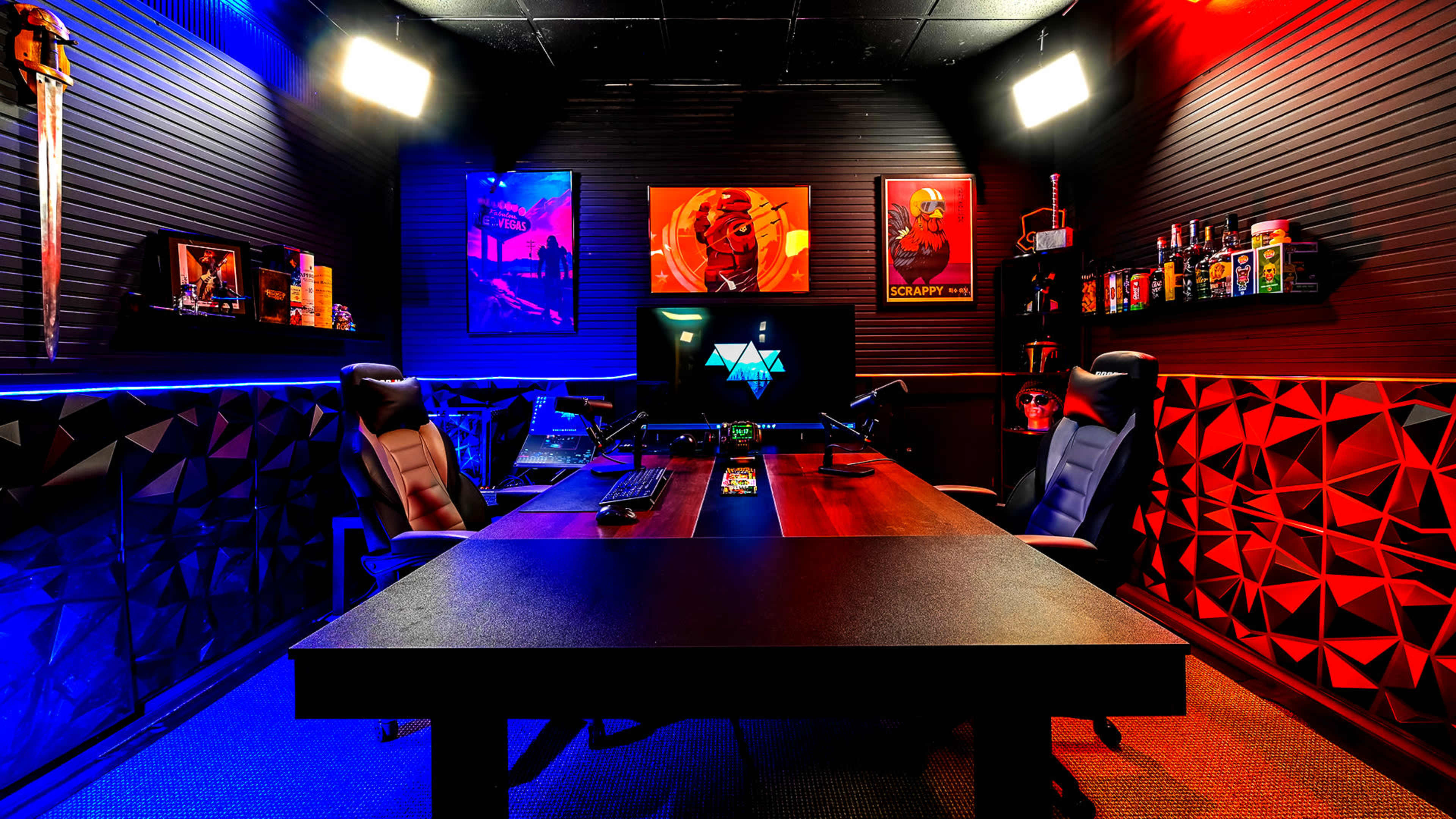 A modern gaming studio features a large table with two gaming chairs, colorful ambient lighting, and framed artwork on the walls.