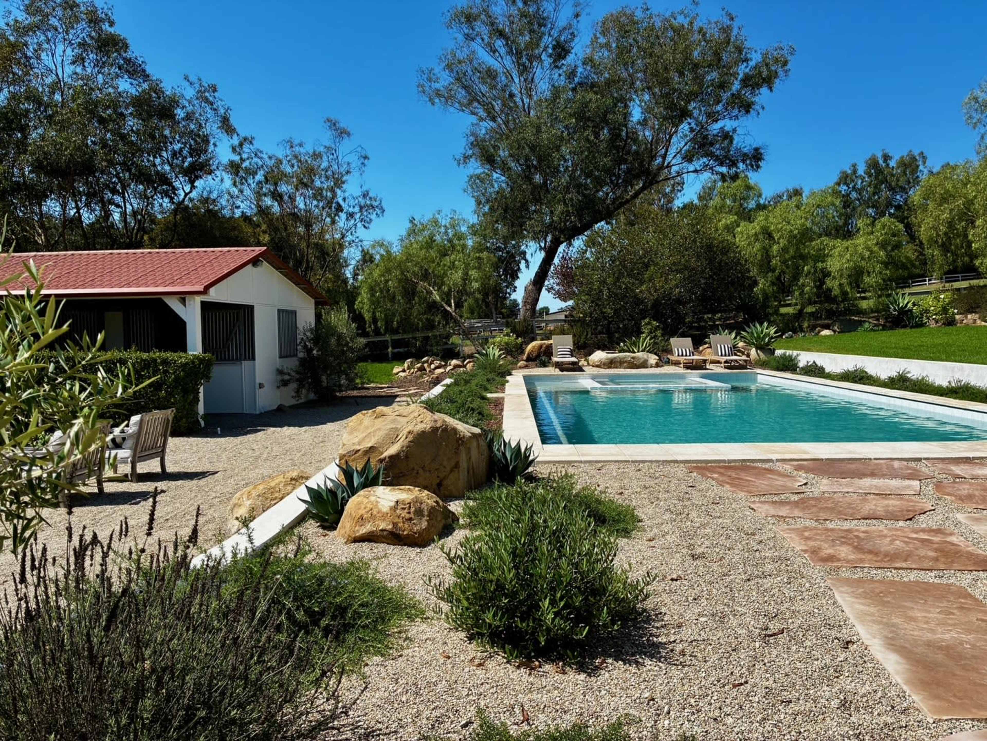 A swimming pool surrounded by rocks and gravel, with lounge chairs and a small shed nearby, against a backdrop of lush greenery and blue skies.