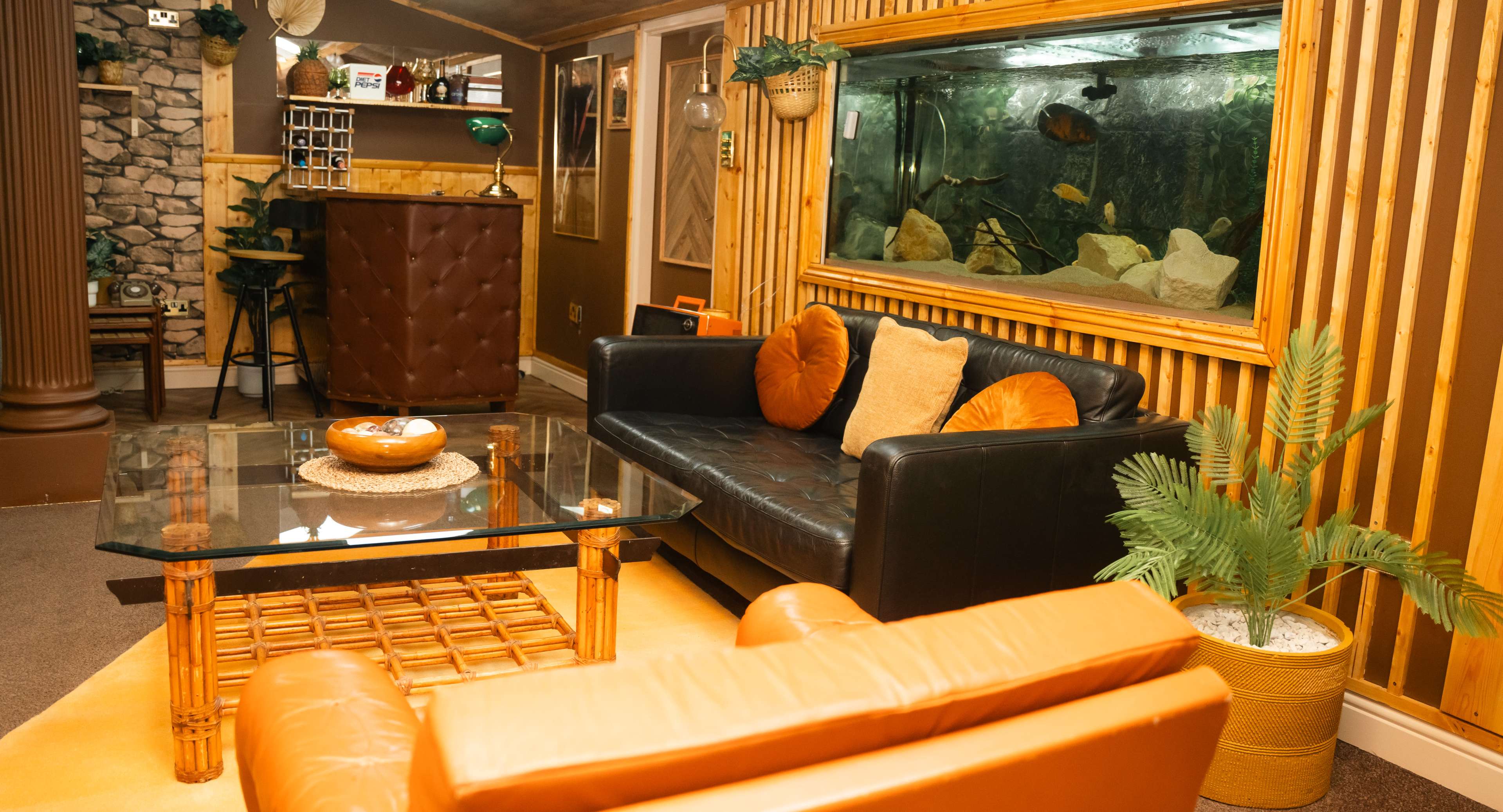 A cozy living room with a black sofa, orange accent cushions, a coffee table, and an aquarium along a wooden-paneled wall.