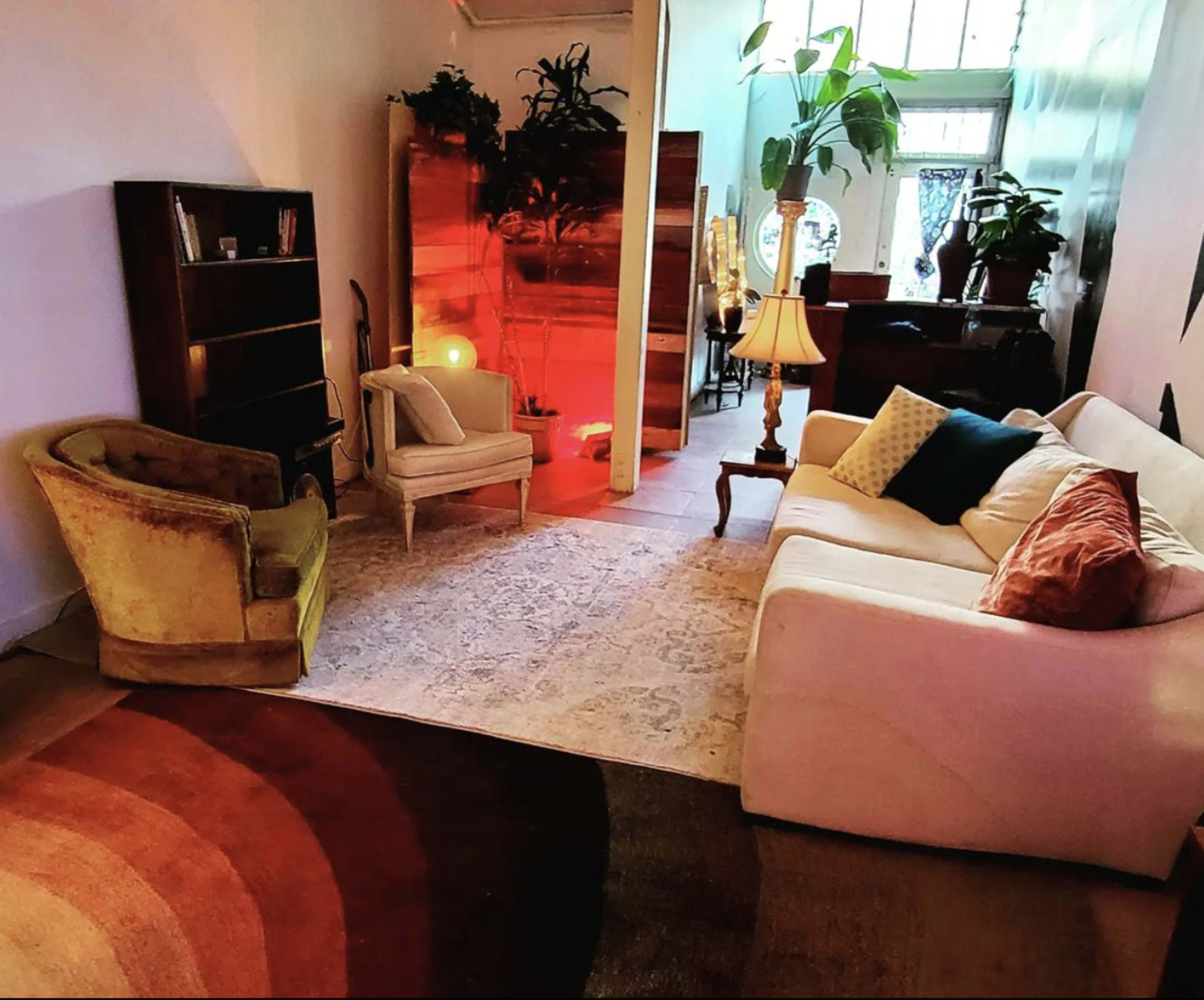 A living room with two armchairs and a sofa arranged around a circular rug, with plants and a bookshelf in the background.