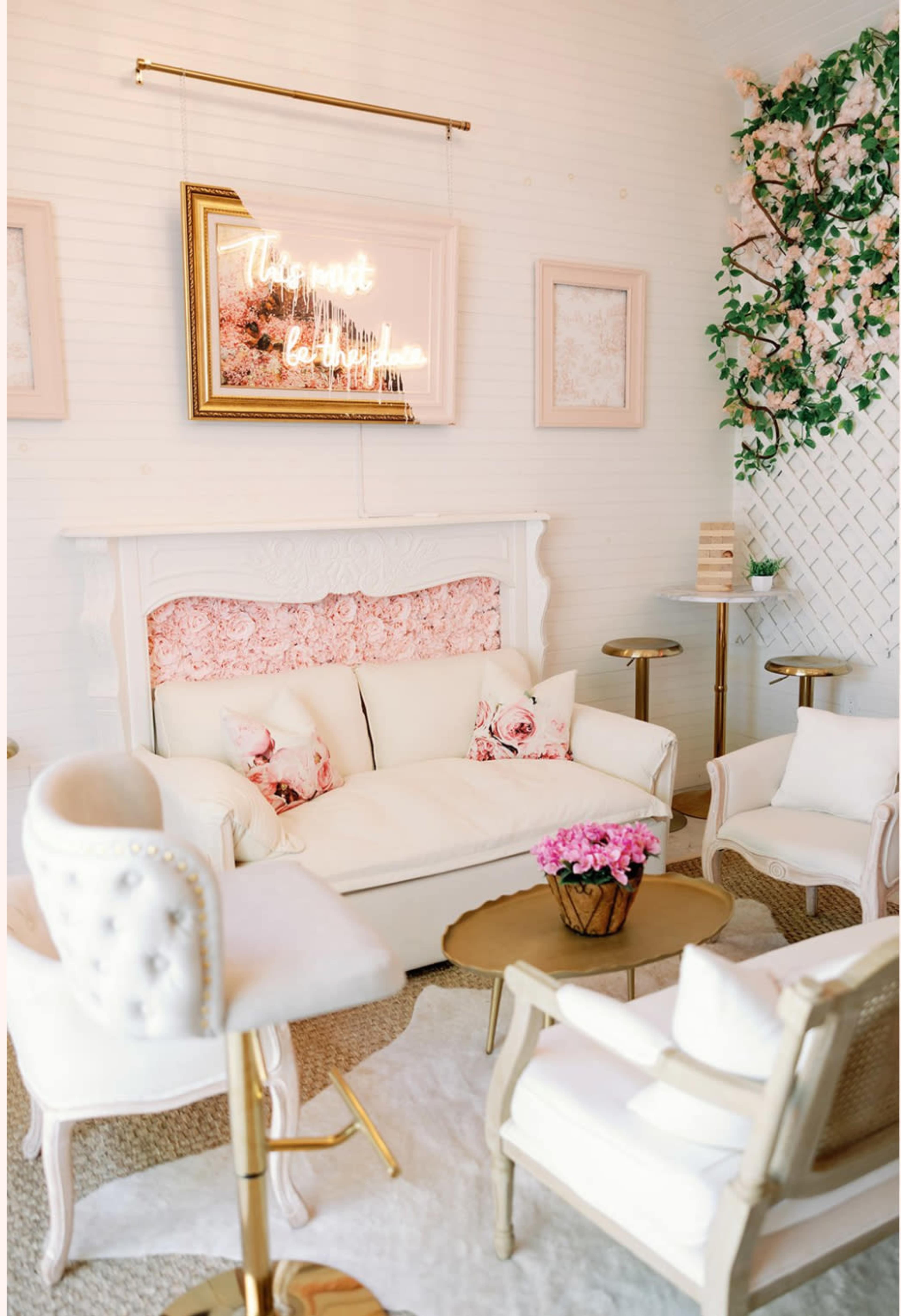 The image depicts a elegantly decorated cozy living room, featuring a white sofa with floral cushions, gold accents, and a neon sign on the wall.
