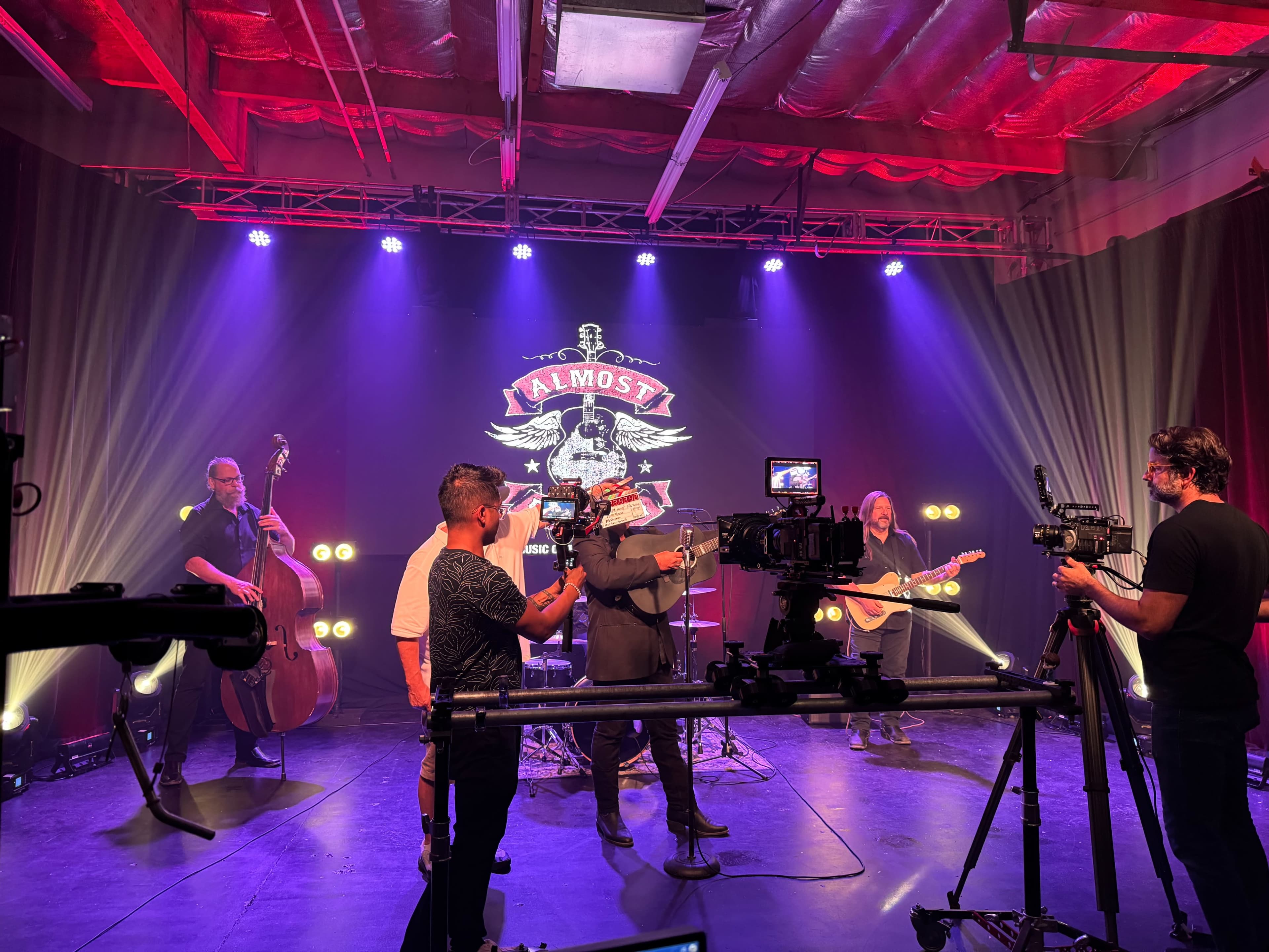 A band performs on stage with multiple cameras capturing the scene, illuminated by colorful lights and a backdrop featuring "Almost."