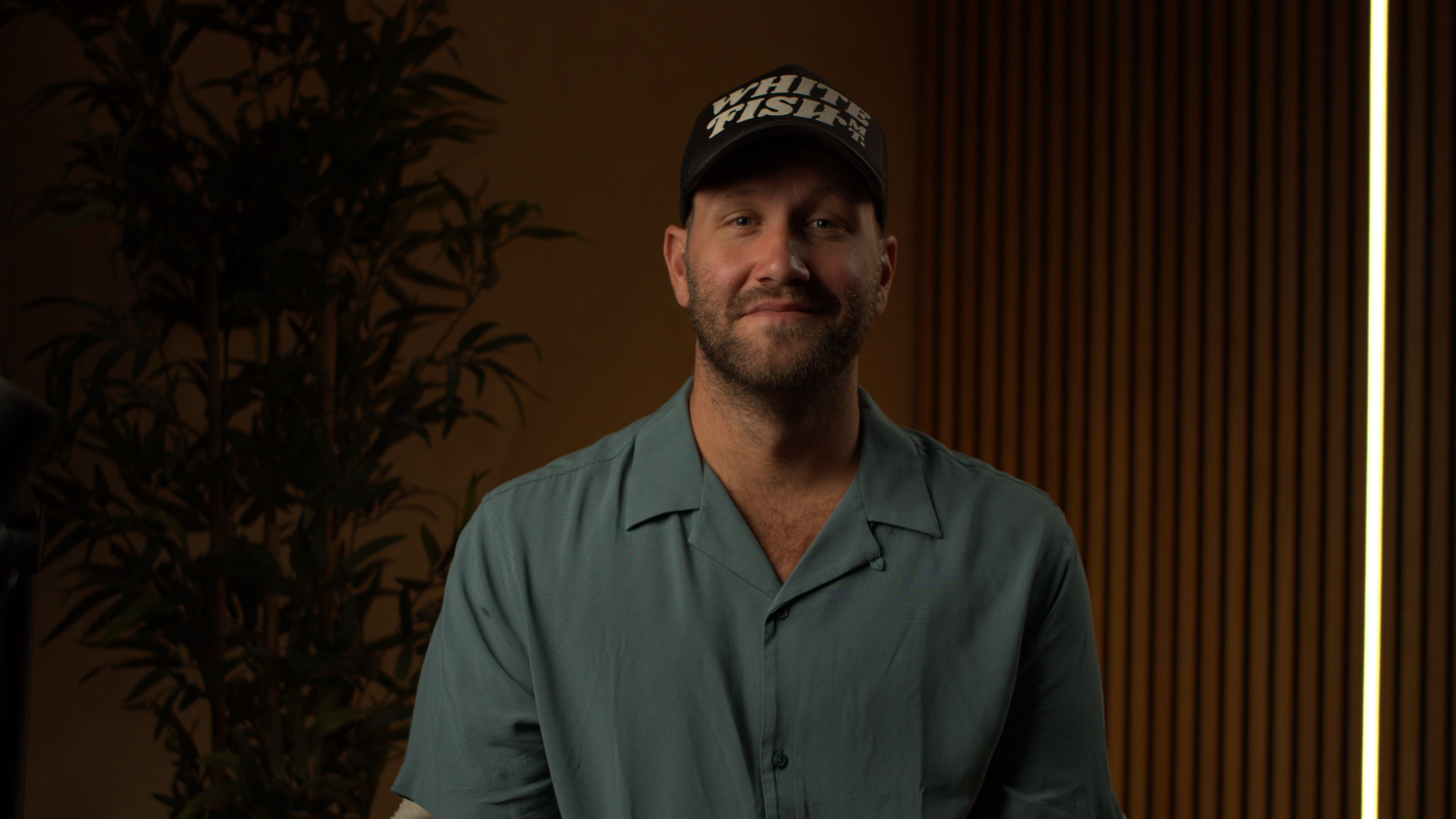 A man wearing a light blue shirt and a black cap sits in front of a plant and a wooden backdrop.