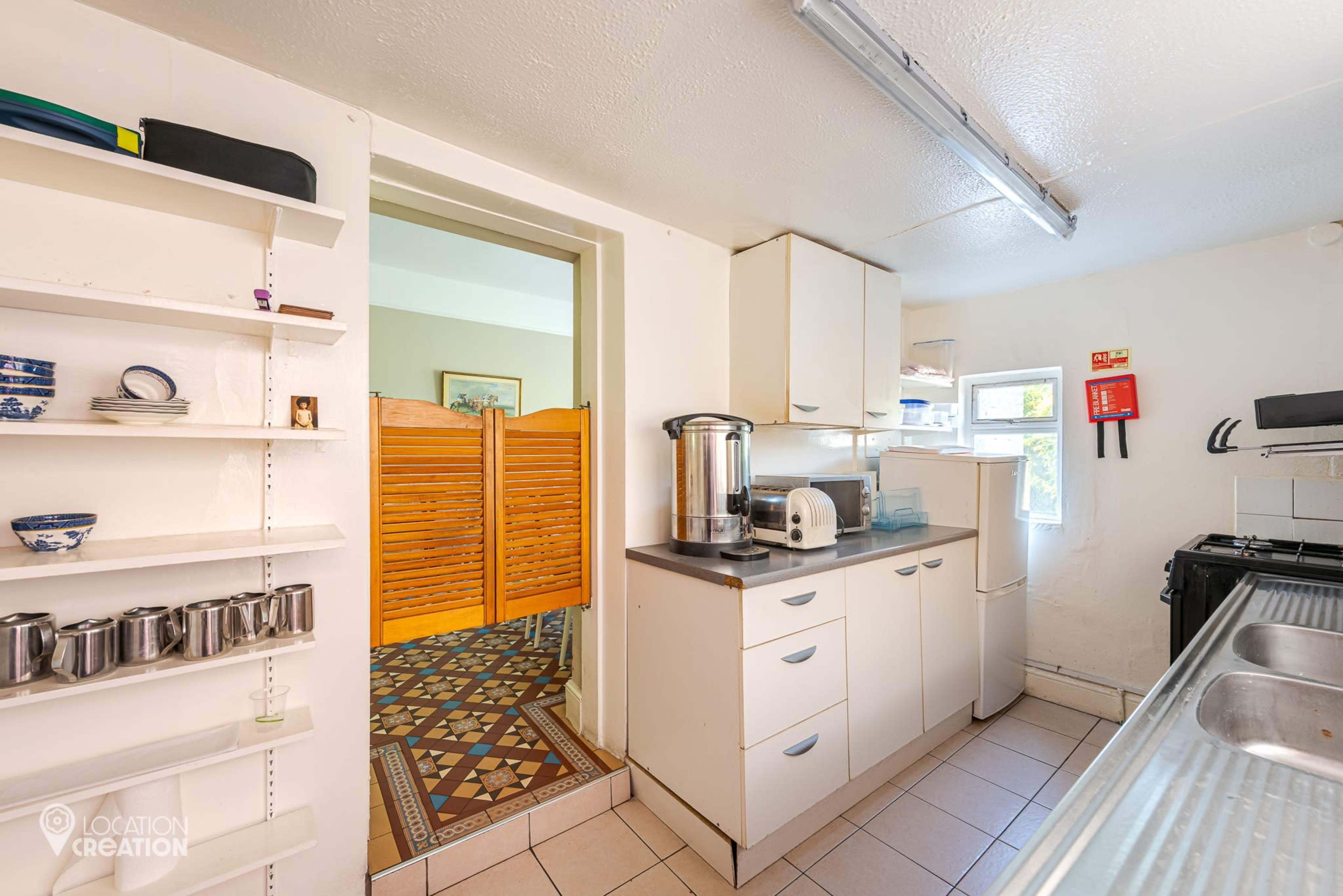 The image shows a kitchen with white cabinets, a stainless steel sink, and shelves displaying cups, adjacent to a hallway leading to another room behind a folding wooden screen.