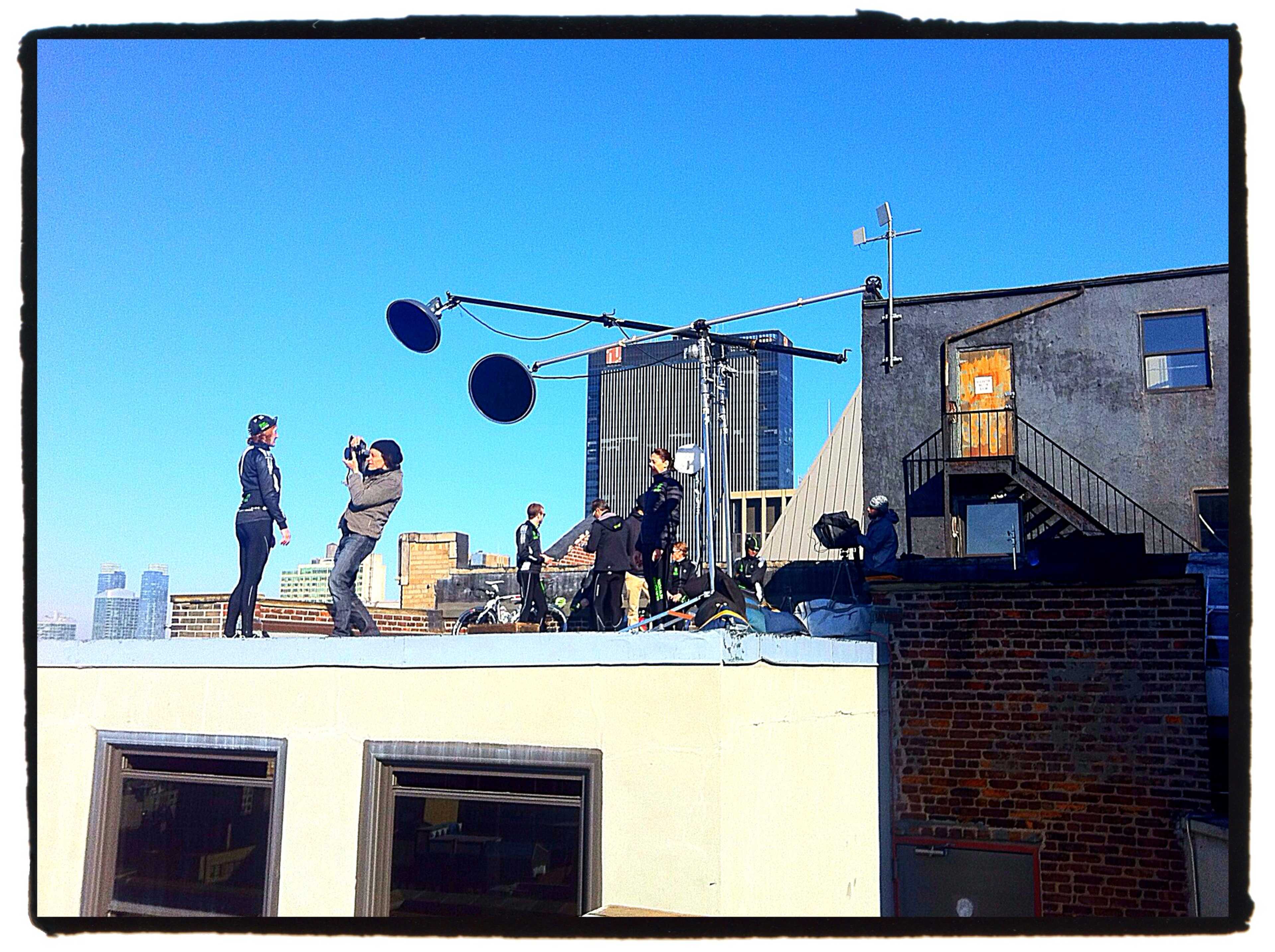 A film crew is shooting a scene on a rooftop with city buildings in the background and equipment set up nearby.